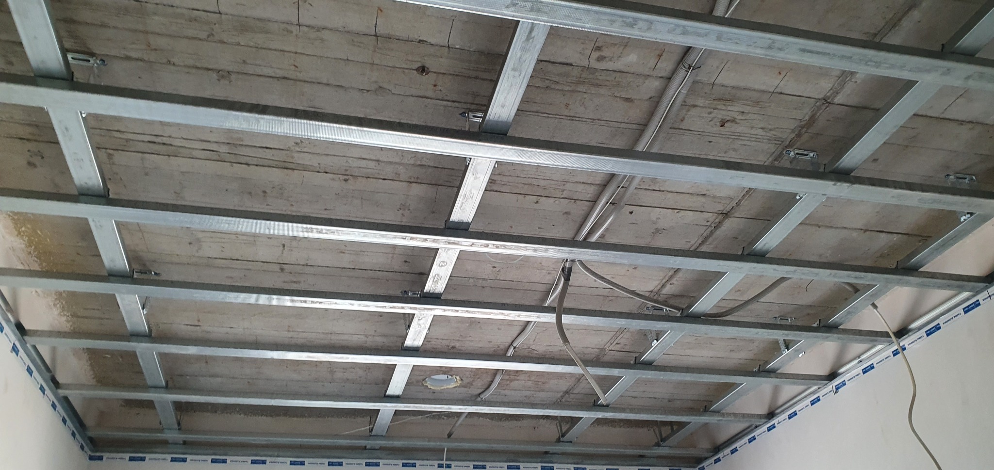 Metalowa konstrukcja podwieszanego sufitu zamontowana pod drewnianym stropem, widoczne przewody elektryczne.
