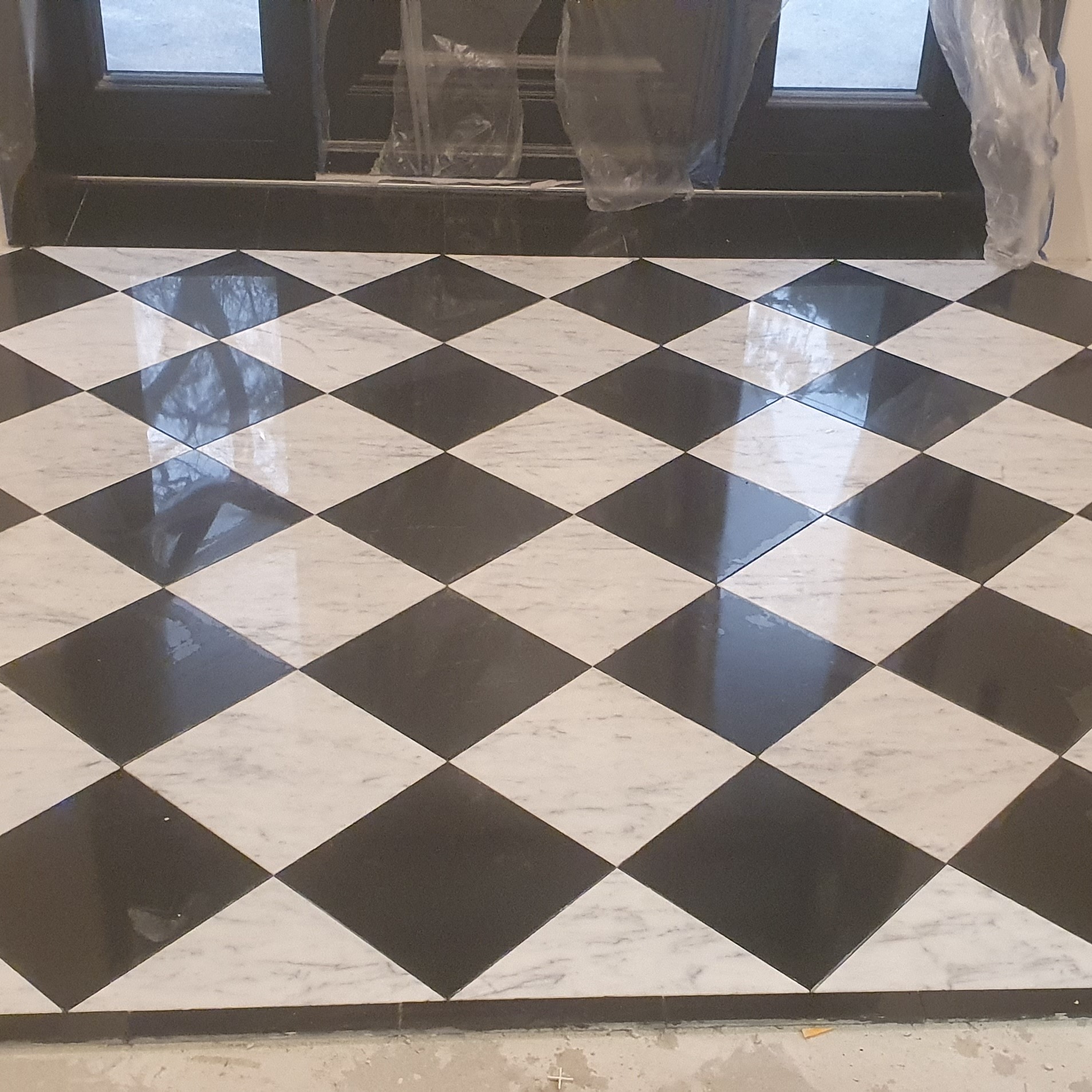 Ułożona podłoga z czarno-białych, błyszczących, marmurowych płytek w romby, widoczne drzwi z folią ochronną, ślady po fugowaniu.