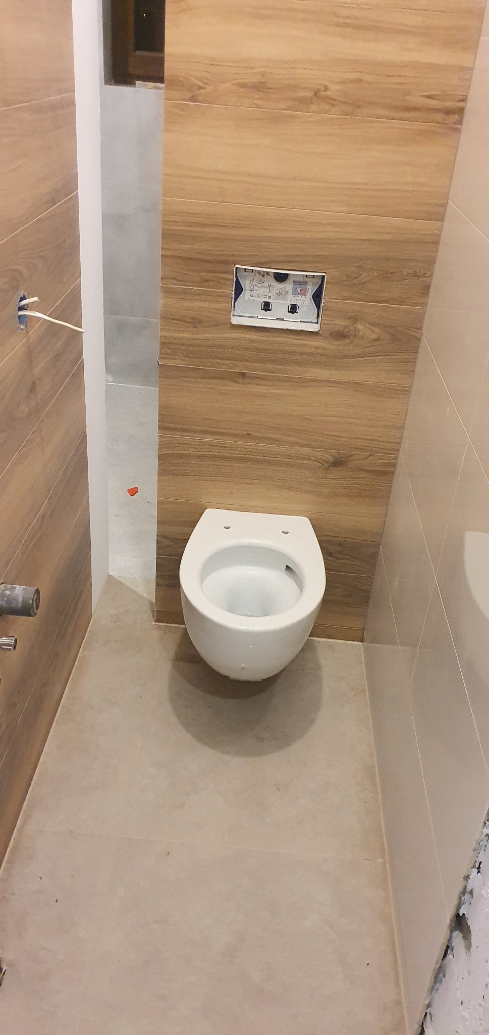 Wisząca miska WC zamontowana na ścianie z panelami imitującymi drewno, widoczna instalacja spłuczki podtynkowej, beżowe płytki podłogowe, częściowo wykończona ściana z prawej strony.