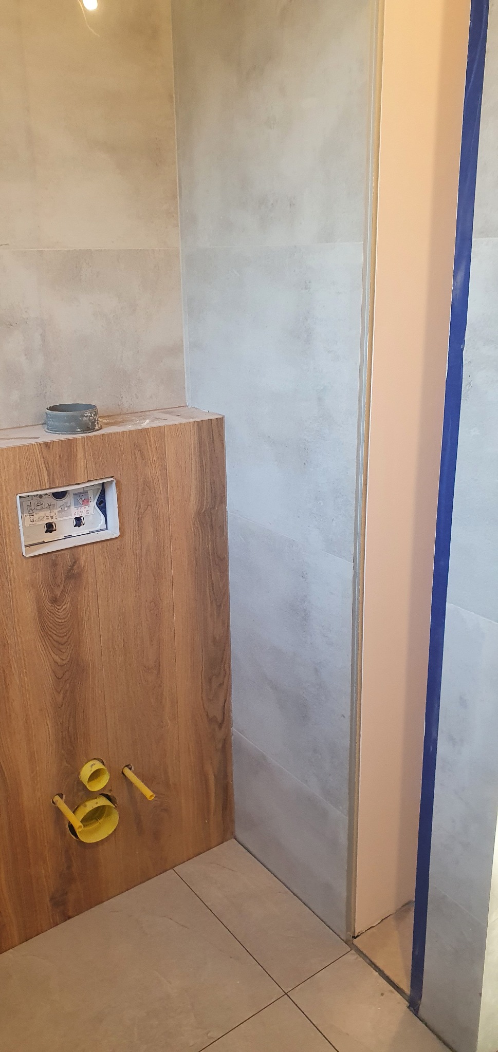 Montaż podtynkowego systemu WC: drewnopodobna obudowa z widocznymi przyłączami kanalizacyjnymi i otwartą puszką instalacyjną, na tle szarych płytek ściennych i podłogowych.