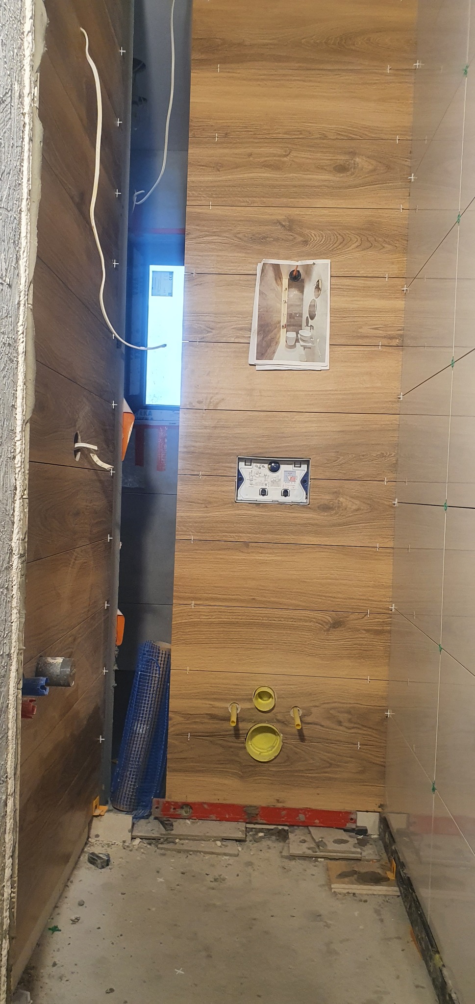 Montaż płytek imitujących drewno na ścianie w łazience, widoczne przyłącza do WC, zamontowany stelaż podtynkowy, poziomica i krzyżyki dystansowe. Na ścianie powieszona kartka z wizualizacją...