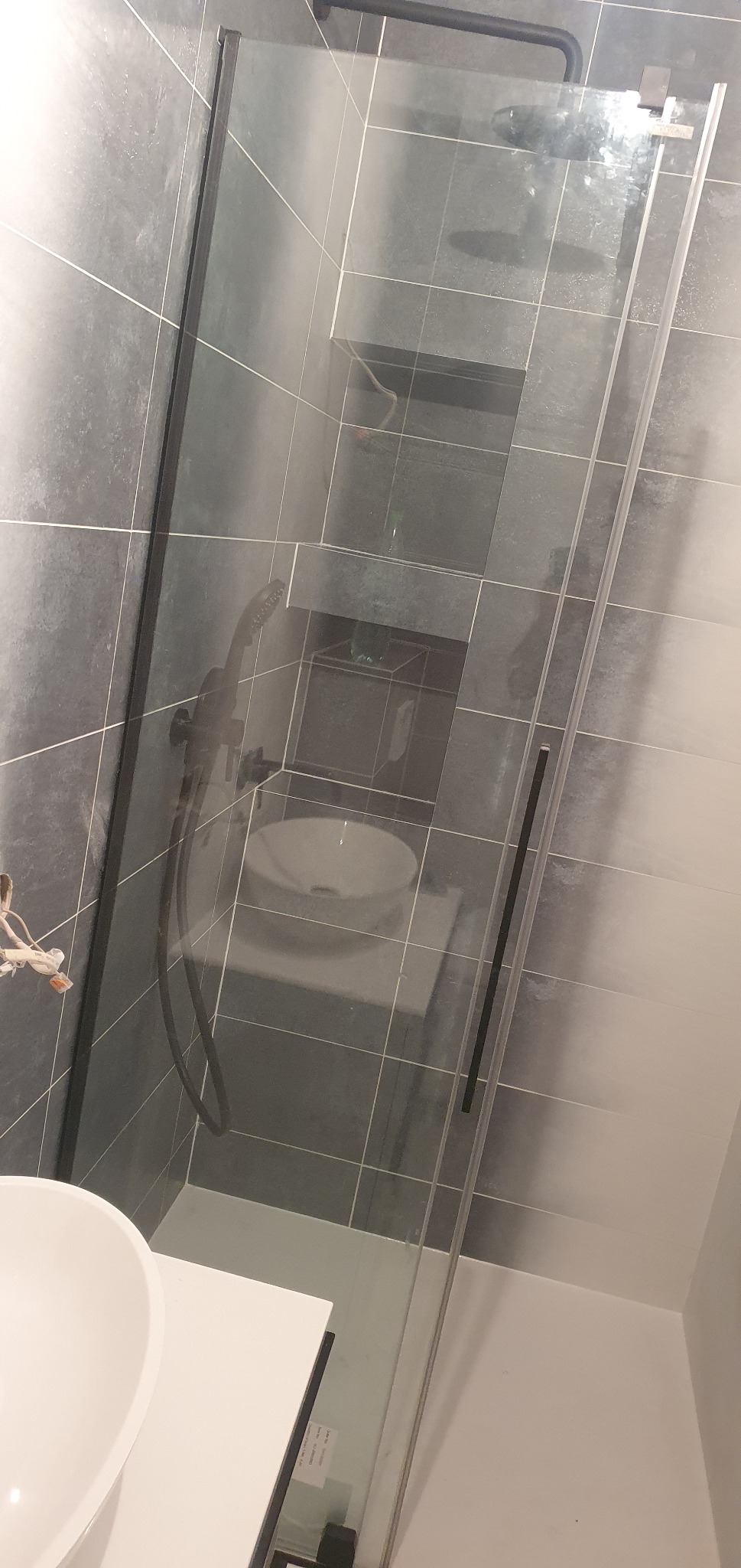 Nowoczesna kabina prysznicowa z czarnymi profilami na tle szarych płytek, widoczna umywalka nablatowa i bateria prysznicowa w kolorze czarnym.