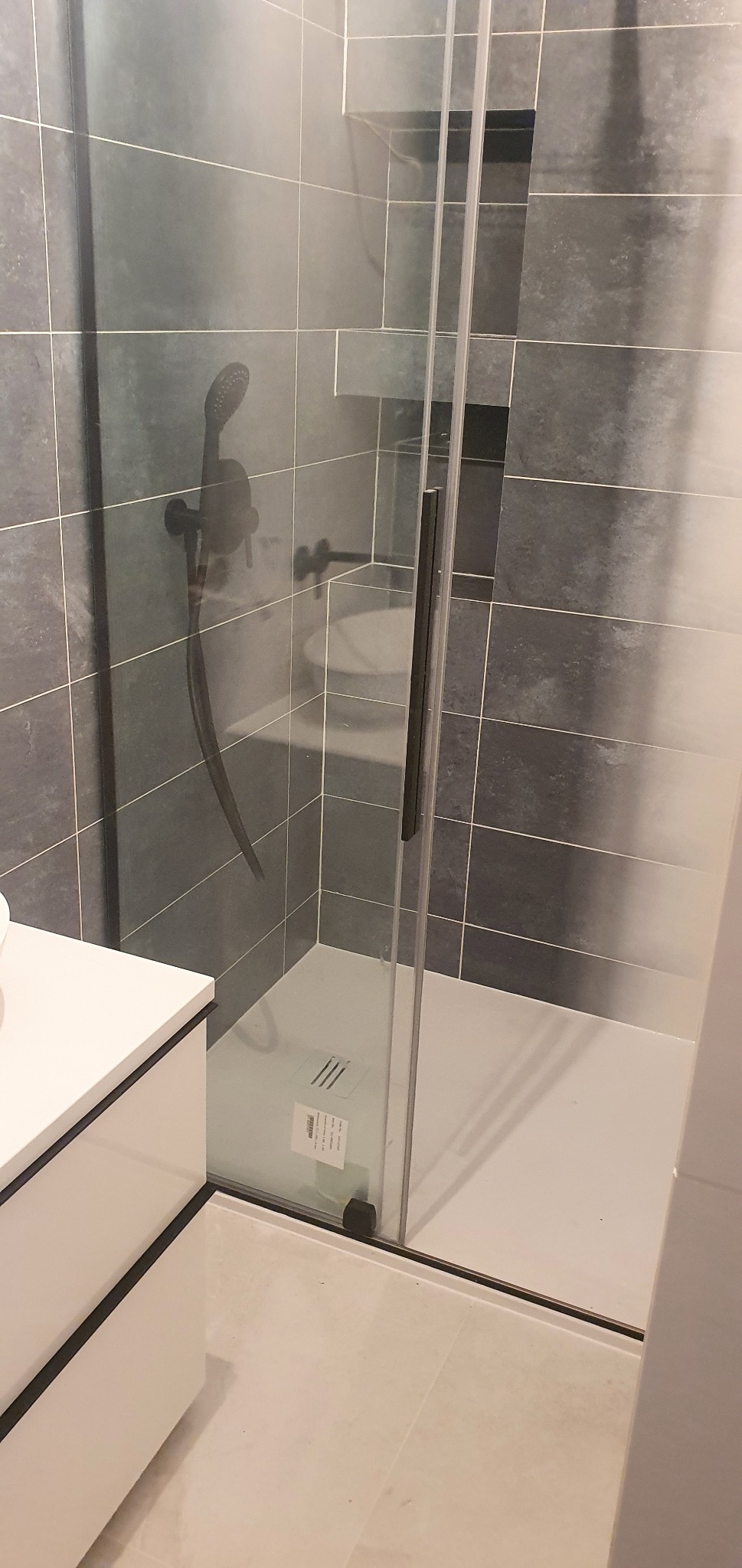 Nowoczesna kabina prysznicowa z czarną armaturą i szarymi płytkami, widoczny fragment białej szafki umywalkowej.