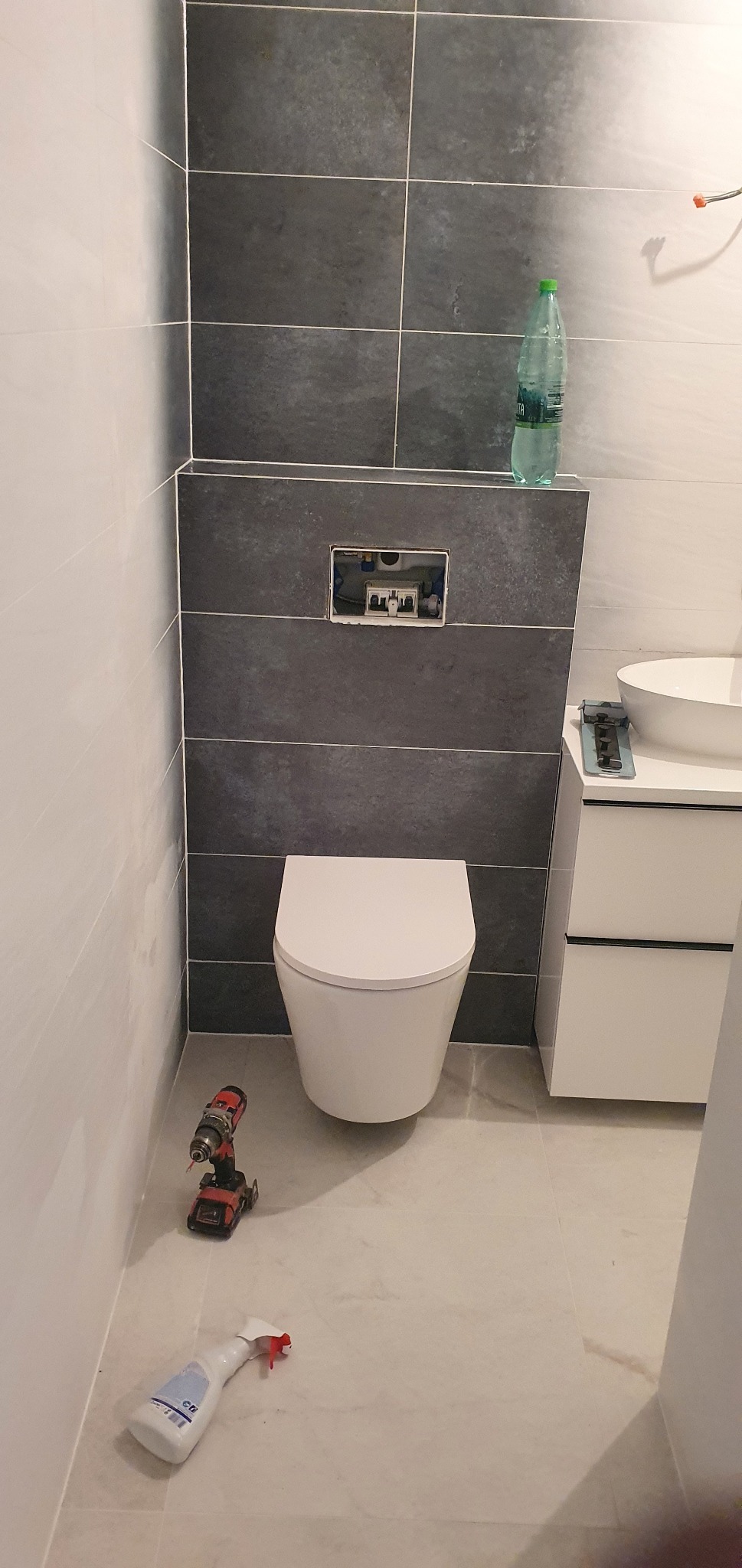 Montaż podtynkowego WC na tle szarych płytek, obok biała szafka z umywalką, na podłodze wiertarka i butelka z płynem do czyszczenia.