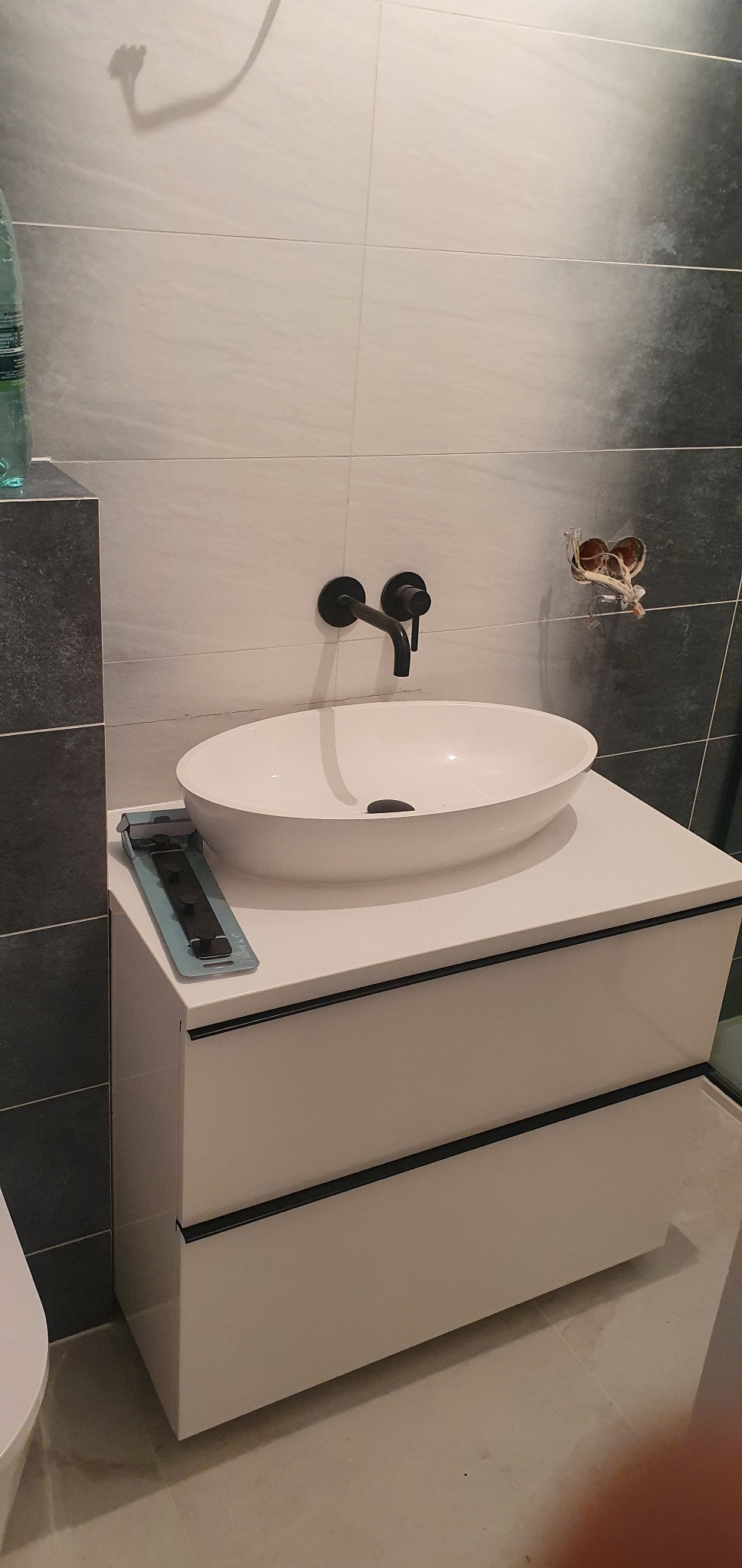 Nowoczesna umywalka nablatowa o owalnym kształcie z czarną armaturą zamontowana na szafce z dwiema szufladami, w łazience wykończonej płytkami w odcieniach szarości i bieli. Widoczne niedokończone...