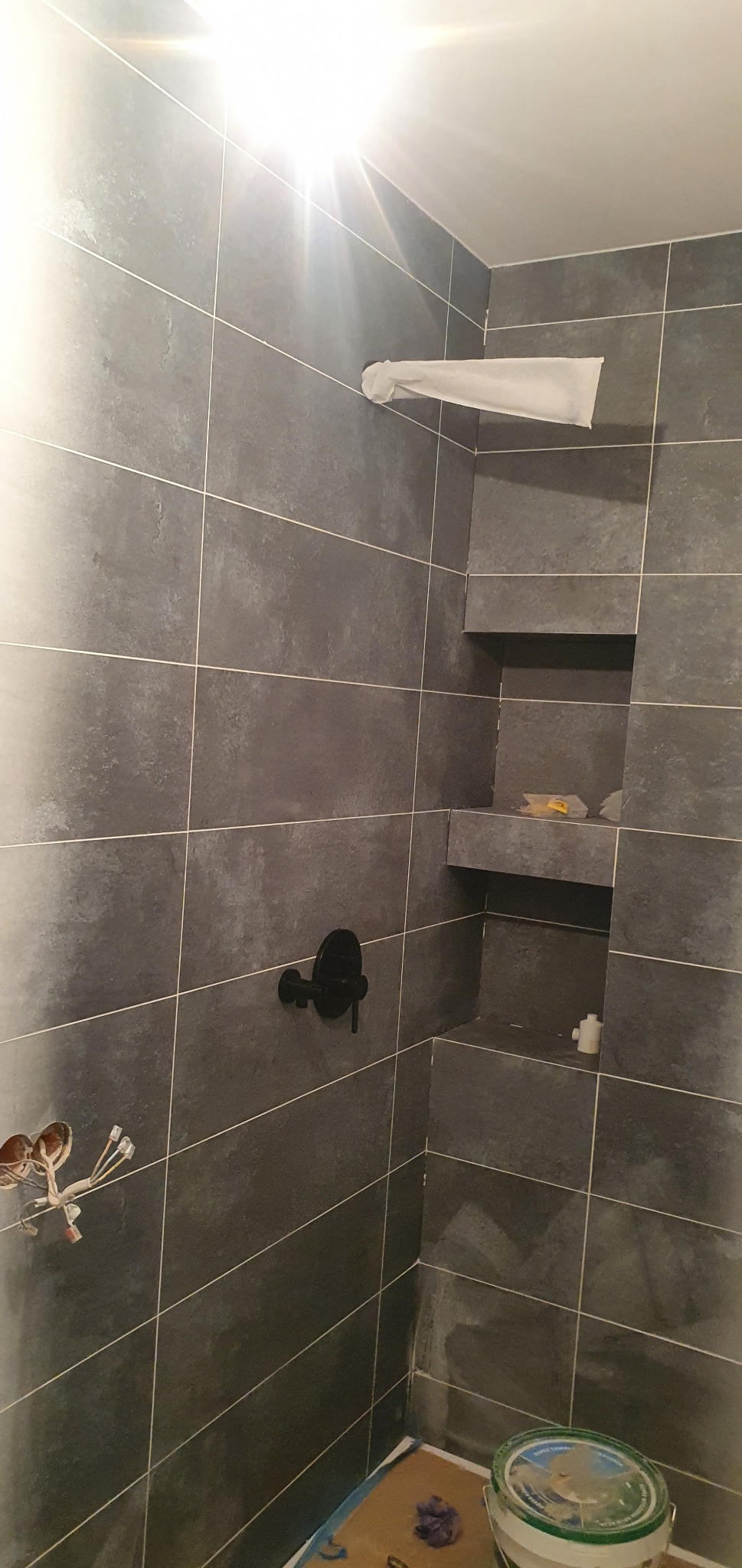 Niedokończona kabina prysznicowa z szarymi płytkami, czarną armaturą i wnękami. Widoczne wystające przewody elektryczne i wiadro na podłodze.