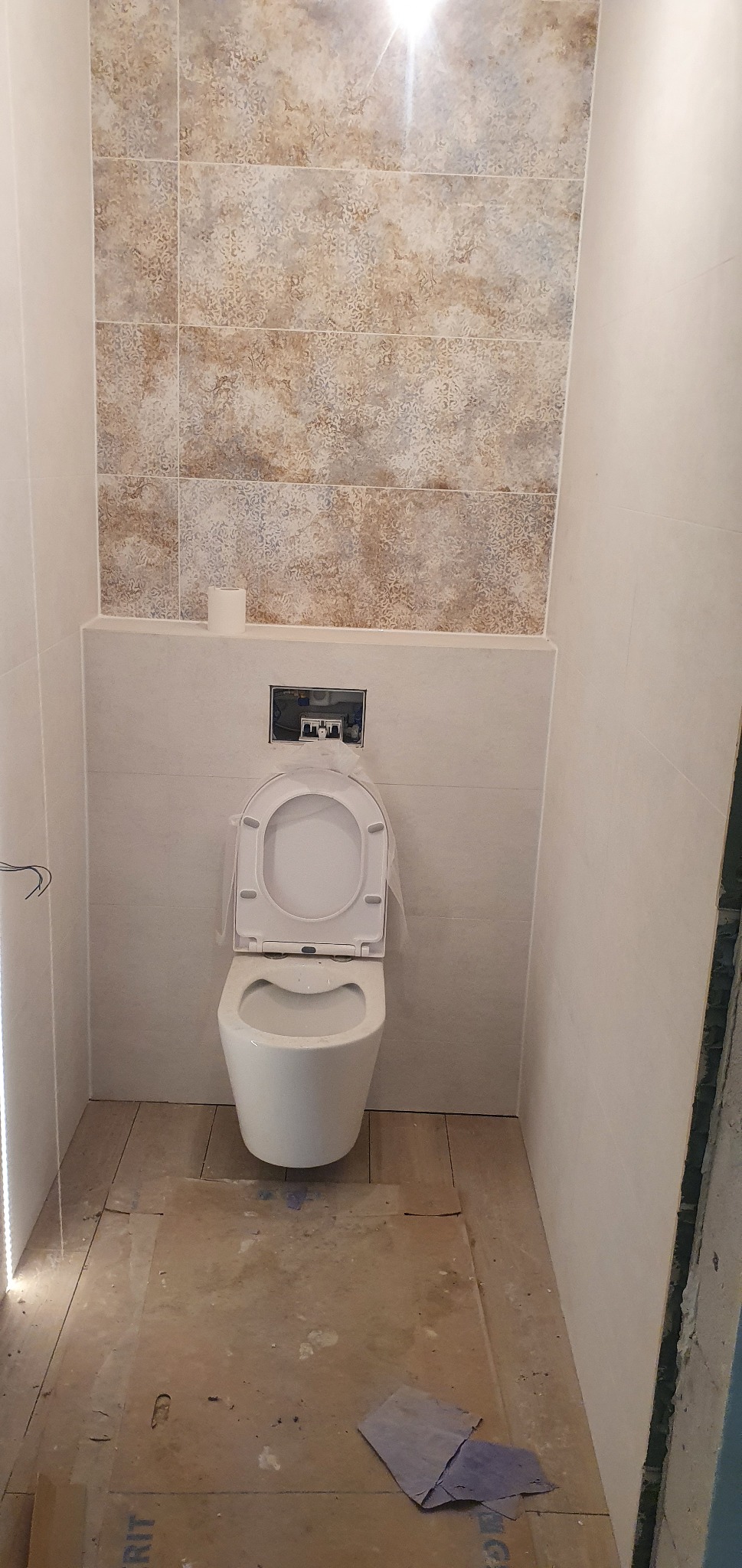 Wisząca toaleta z otwartą klapą, zamontowana na tle beżowych płytek z wzorem, z widoczną instalacją i rolką papieru toaletowego, podłoga zabezpieczona kartonem.