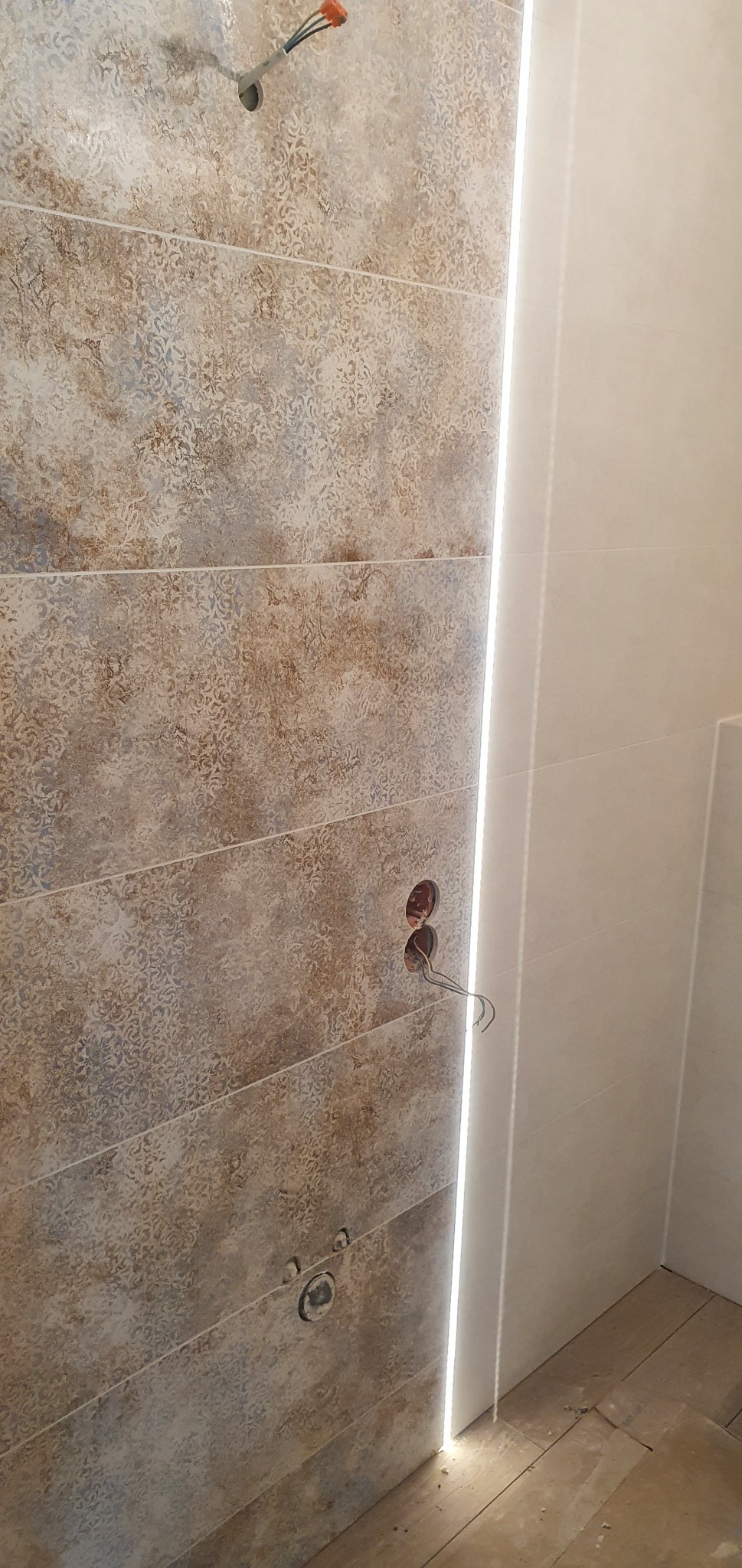 Ściana łazienki wyłożona płytkami w odcieniach beżu i brązu z delikatnym wzorem, oświetlona pionową listwą LED, widoczne otwory na instalacje elektryczne i wodne z wystającymi przewodami.