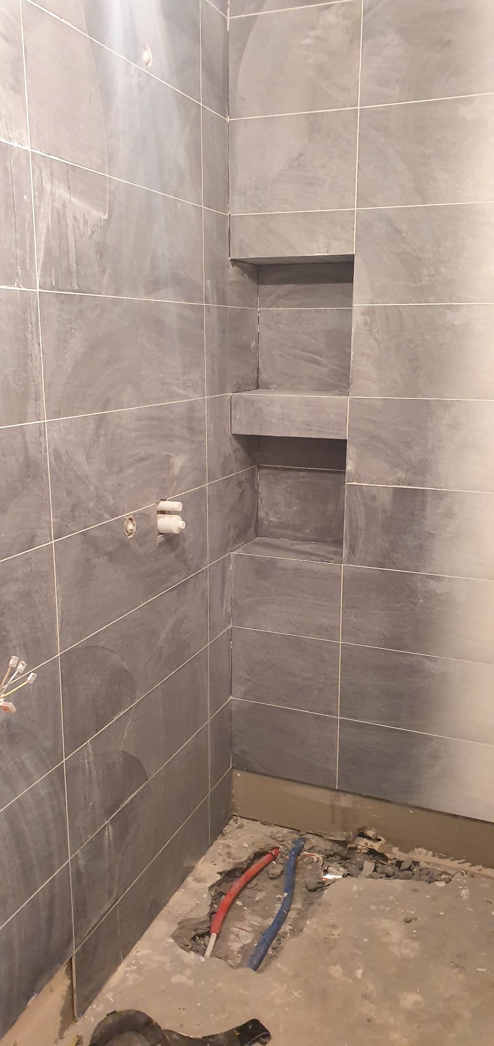 Wykończenie łazienki w trakcie: szare, matowe płytki ceramiczne na ścianach kabiny prysznicowej z wnękami na kosmetyki, widoczne rury instalacji wodnej w podłodze przed położeniem posadzki.