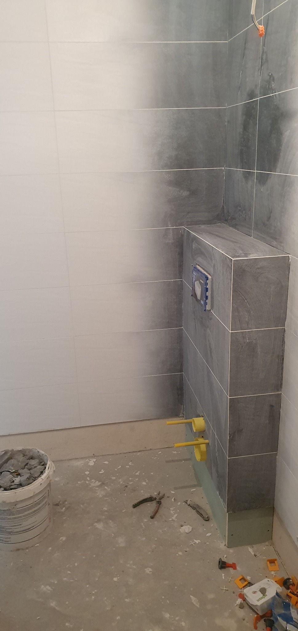 Wnętrze łazienki w trakcie remontu: częściowo wyłożone szarymi i białymi płytkami ściany, widoczna instalacja podtynkowego WC z żółtymi przyłączami, obok wiadro z resztkami płytek i narzędzia...