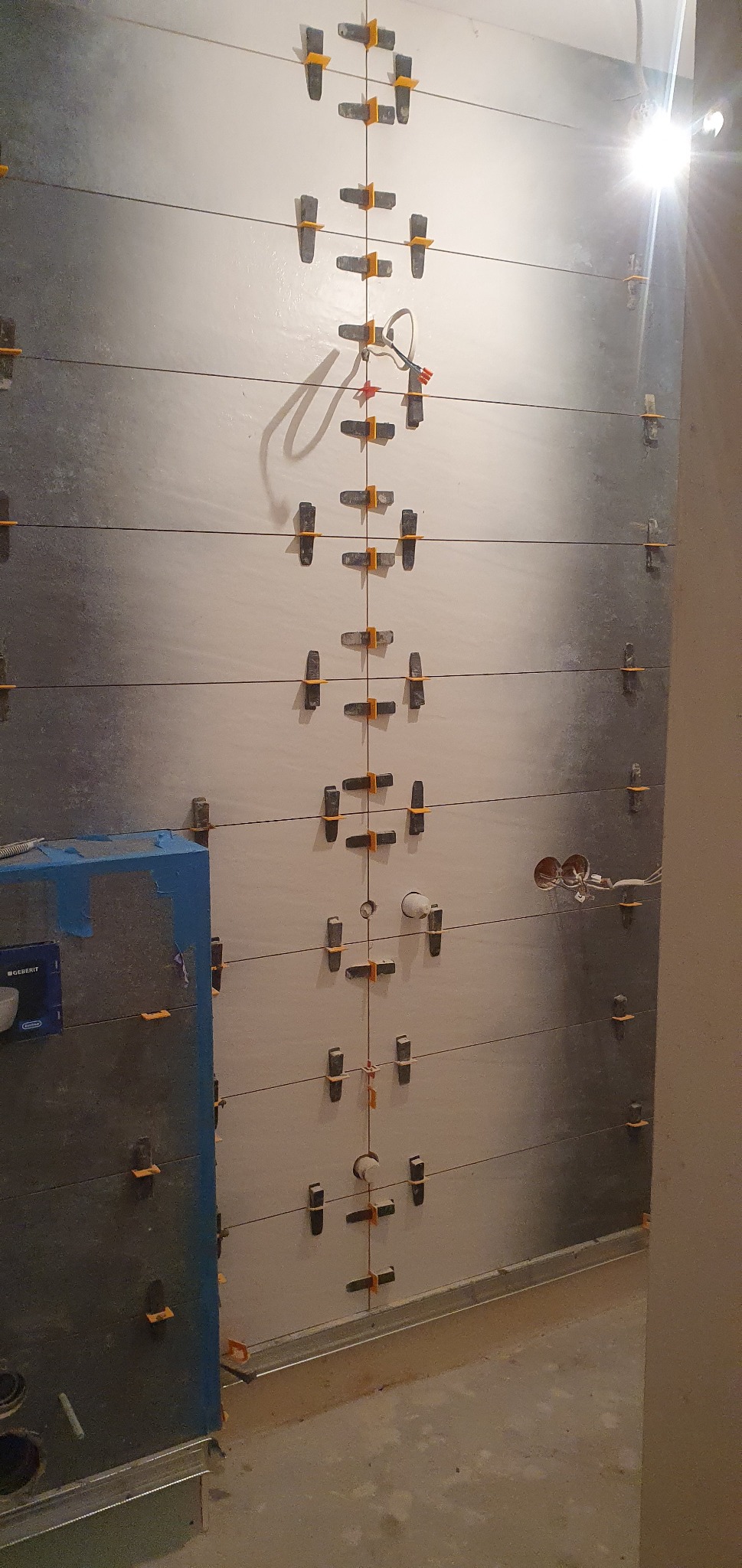 Ściana w trakcie układania płytek, z widocznymi systemami poziomowania płytek i wystającymi przewodami elektrycznymi.