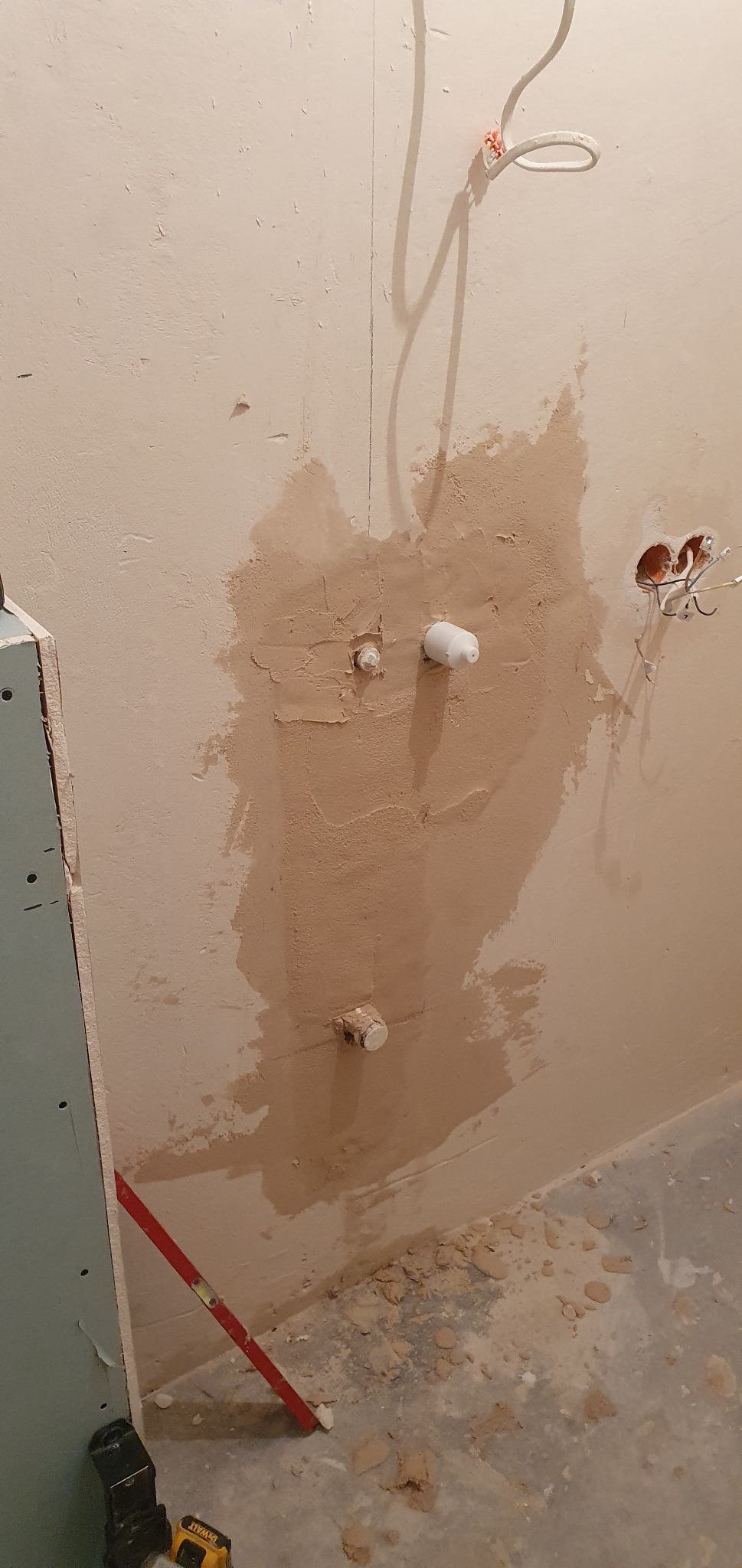 Fragment ściany w trakcie prac wykończeniowych: widoczne wystające rury, zakryte świeżym tynkiem, oraz niezabezpieczone przewody elektryczne i poziomica oparta o płytę karton gips.