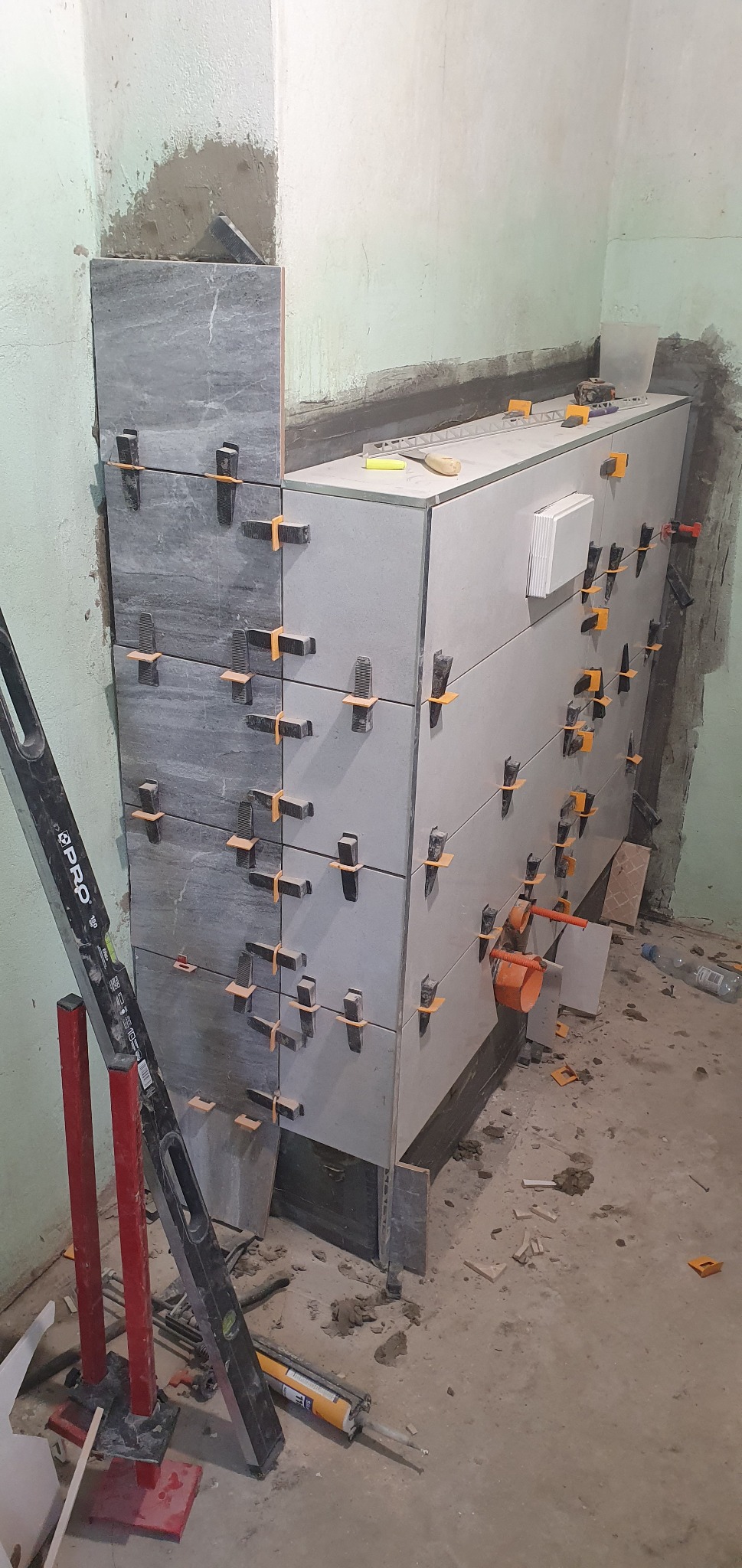 Proces układania szarych i białych płytek ceramicznych na ścianie, z użyciem klipsów poziomujących i poziomicy PRO, widoczne fragmenty instalacji hydraulicznej