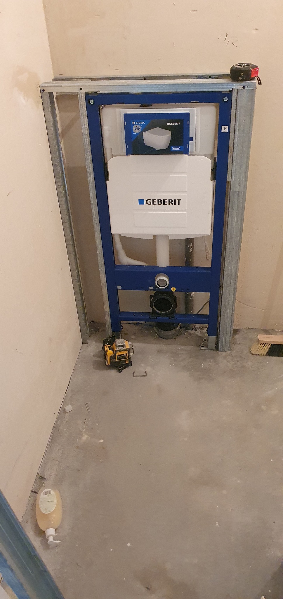 Stelaż podtynkowy Geberit Sigma zamontowany w łazience, widoczny laser krzyżowy DeWalt, miarka zwinięta na stelażu i butelka z płynem do mycia podłóg