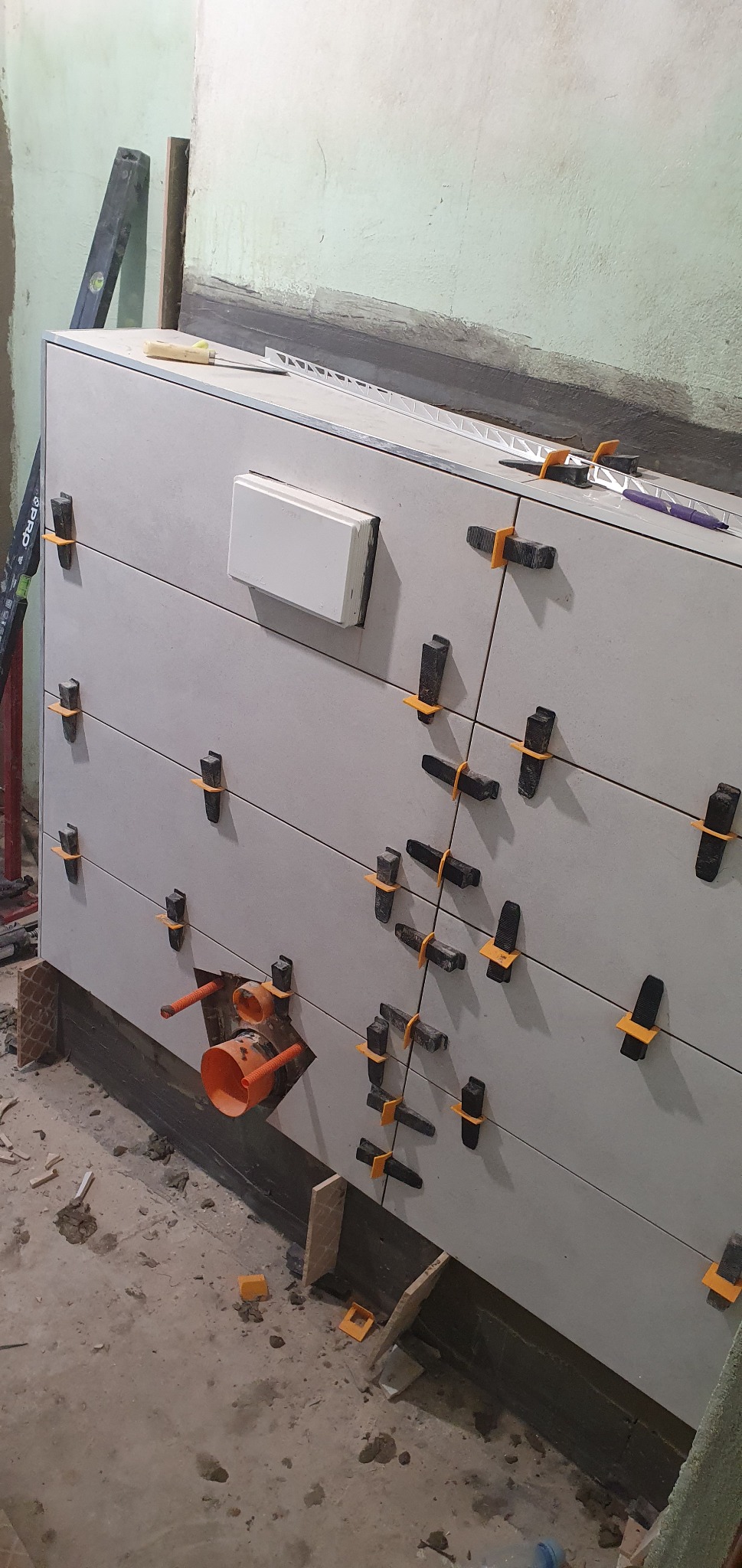 Świeżo ułożone, szare płytki ścienne z pomarańczowymi elementami poziomującymi i czarnymi klinami, widoczne rury kanalizacyjne w ścianie, poziomica i narzędzia na górnej krawędzi płytek, surowy...