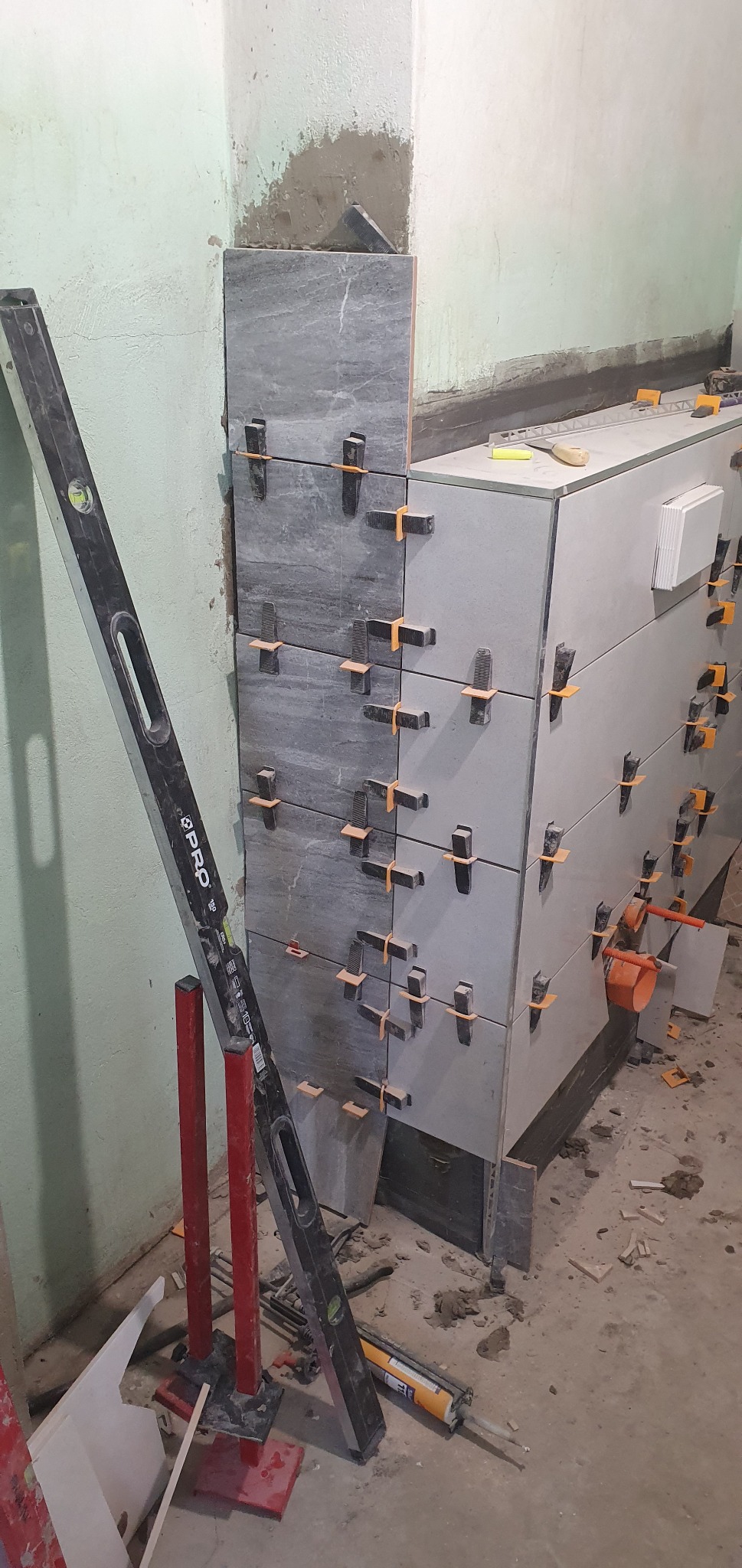 Układanie płytek na ścianie, widoczne poziomica, krzyżyki dystansowe i fragment ściany z pozostałościami zaprawy.