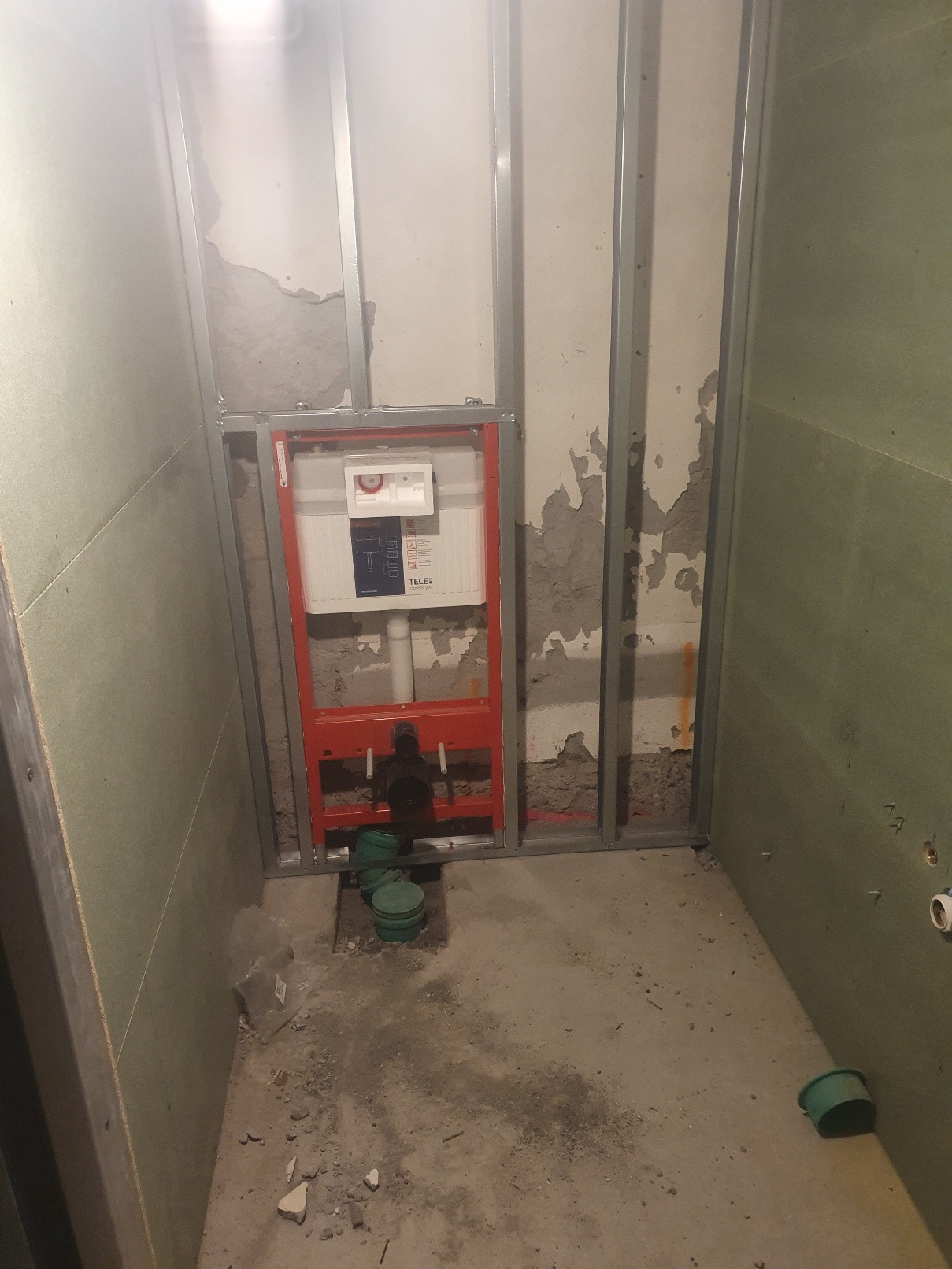 Stelaż podtynkowy WC marki TECE zamontowany w konstrukcji z profili stalowych, widoczne fragmenty instalacji wodno-kanalizacyjnej i surowa ściana z ubytkami tynku.