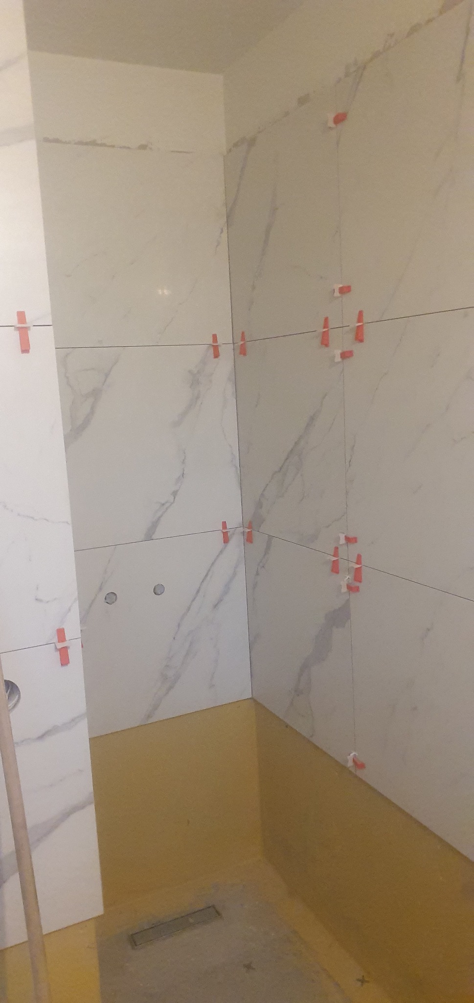 Wnętrze łazienki w trakcie układania białych płytek imitujących marmur, z użyciem czerwonych krzyżyków dystansowych, widoczna żółta hydroizolacja podłoża.
