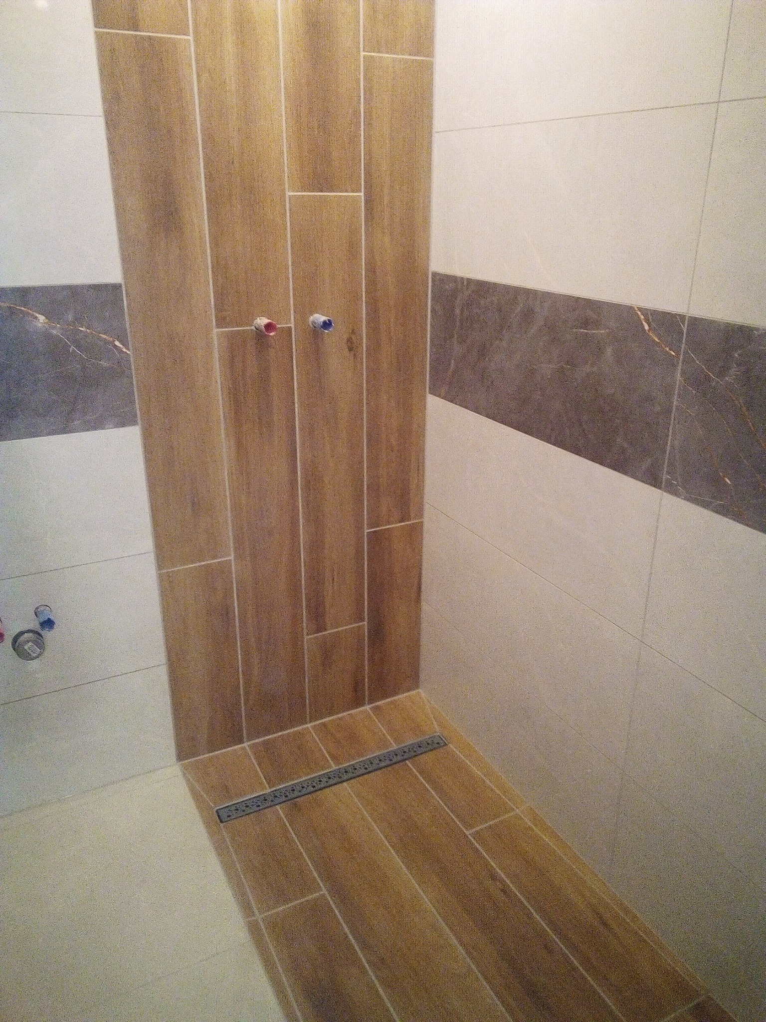 Wykończona kabina prysznicowa z drewnopodobnymi płytkami w odcieniach brązu, z widocznym odpływem liniowym i punktami instalacji wodnej z czerwonym i niebieskim oznaczeniem.