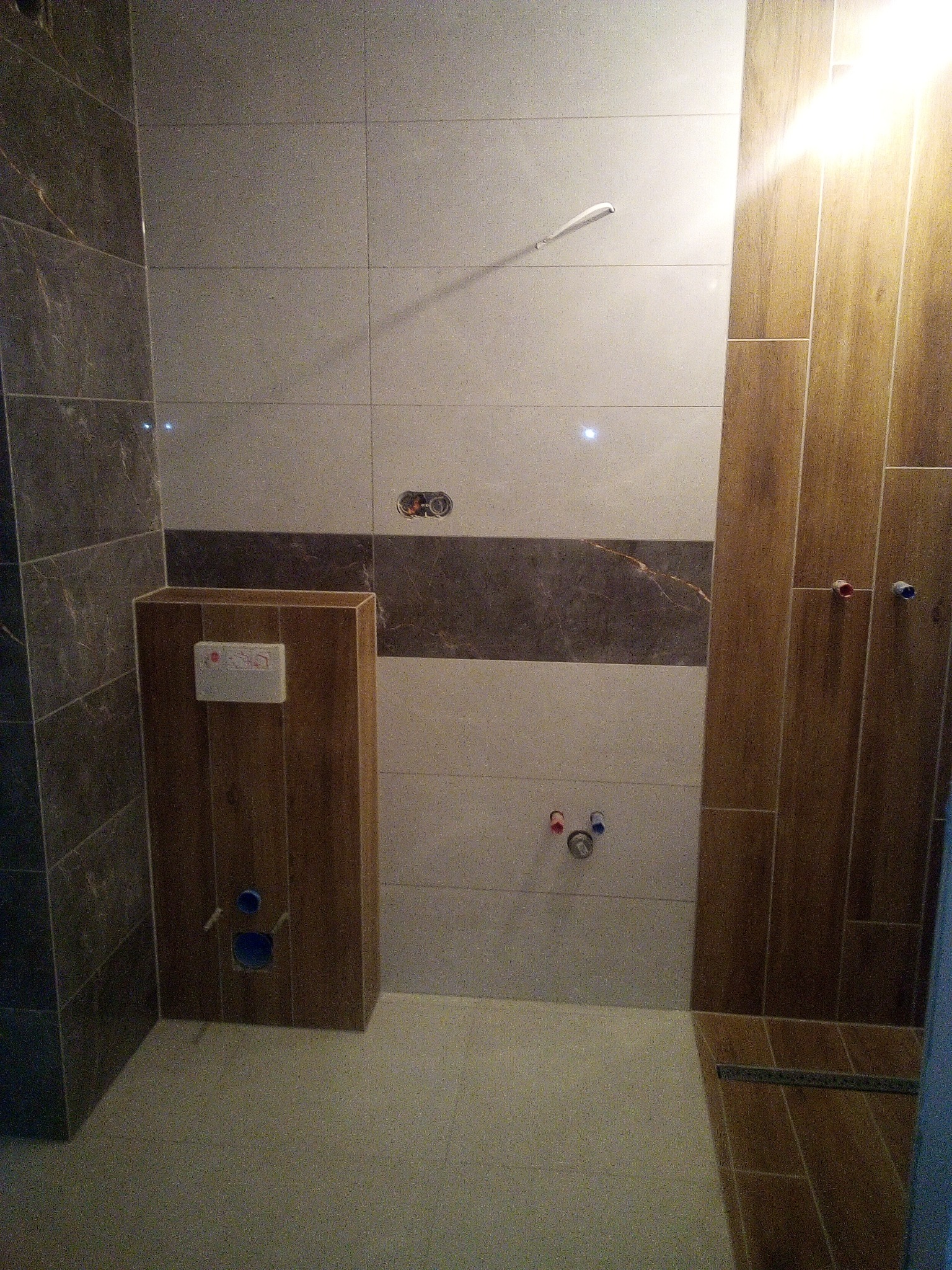 Wykończona łazienka z brązowo-beżowymi płytkami, zabudowa WC z drewnopodobnych płytek, widoczne przyłącza wodne i odpływ. Świeżo położona glazura.