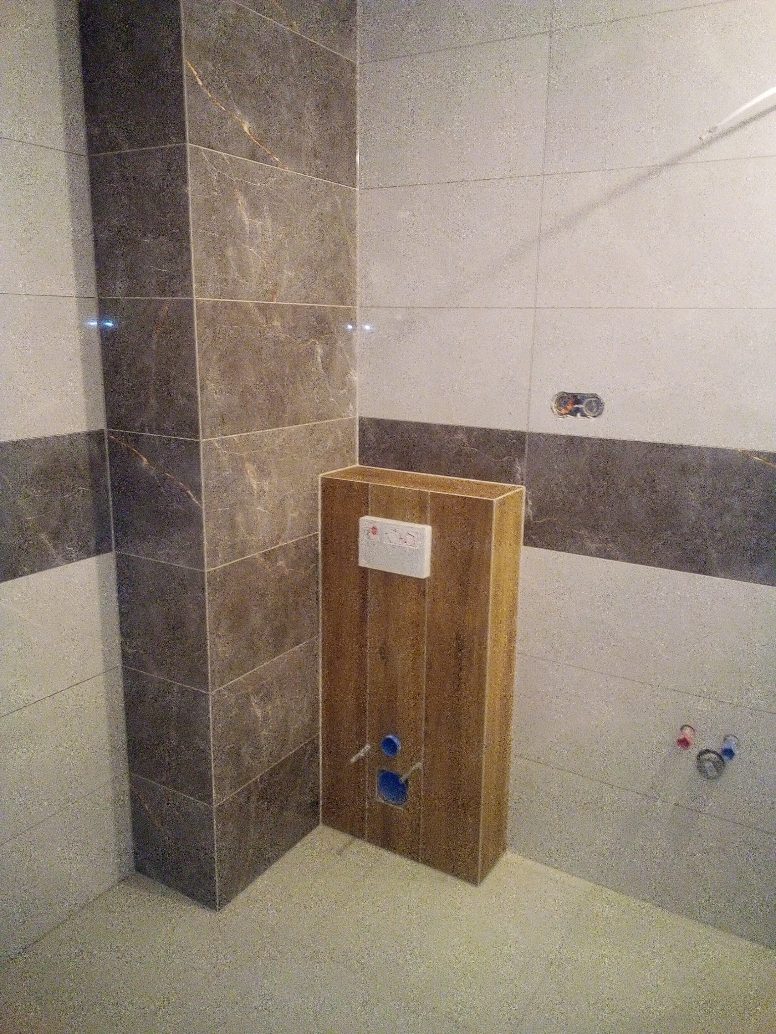 Wykończona łazienka z brązowo-beżowymi płytkami imitującymi marmur, zabudowany stelaż WC z drewnianą obudową i białym przyciskiem, widoczne przyłącza wodne.