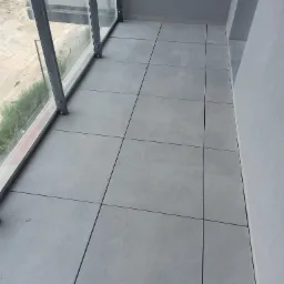 Szare, kwadratowe płytki na balkonie z szarą ścianą i szklaną balustradą z metalowymi ramami.