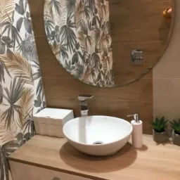 Nowoczesna łazienka z umywalką nablatową w kształcie misy, baterią chromowaną, drewnianym blatem i lustrem w okrągłej ramie, odbijającym tapetę w liście.