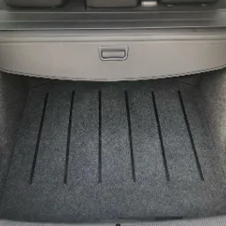 Czysty bagażnik samochodu z szarą wykładziną i czarnymi listwami ochronnymi, zasłonięty roletą.