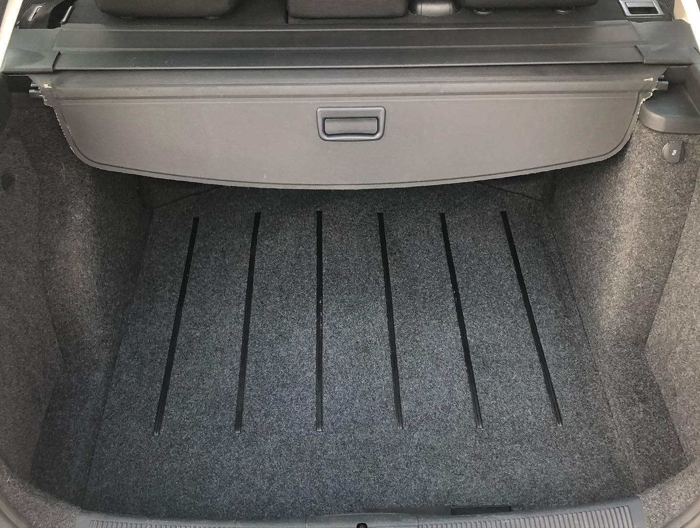 Czysty bagażnik samochodu z szarą wykładziną i czarnymi listwami ochronnymi, zasłonięty roletą.