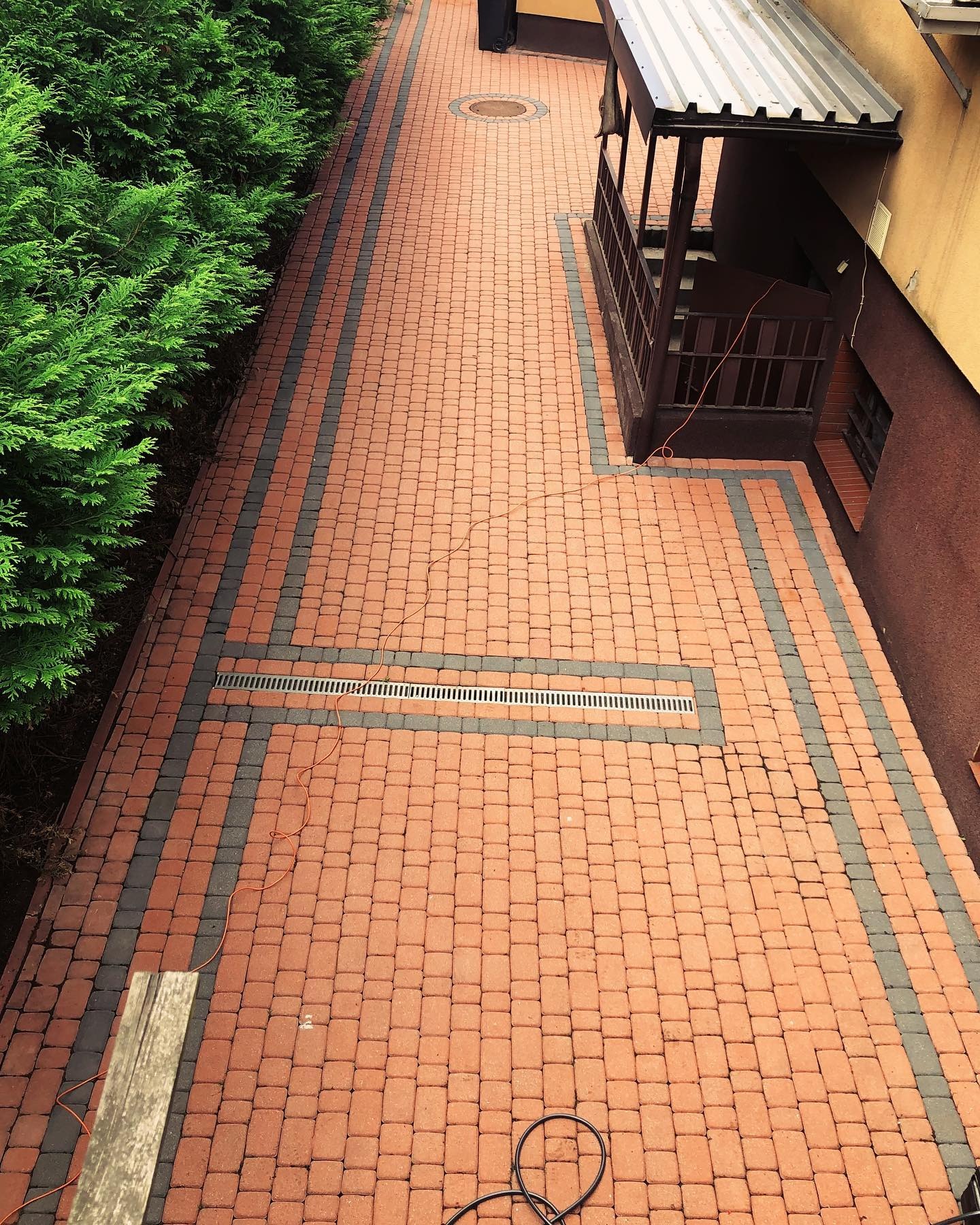 Ułożona kostka brukowa w odcieniach czerwieni i szarości, z widocznym odpływem liniowym, przylegająca do budynku z zadaszonym wejściem i zielonymi krzewami, sfotografowana z góry.
