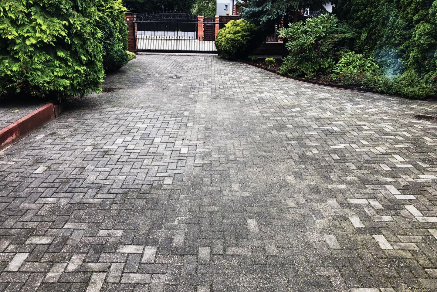 Szeroki kadr na szarą kostkę brukową ułożoną we wzór jodełki, prowadzącą w kierunku czarnej bramy wjazdowej z czerwonymi ceglanymi słupkami, otoczoną bujną zielenią.