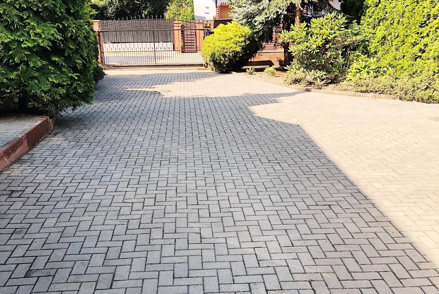 Ułożona szara kostka brukowa prowadząca do bramy wjazdowej, otoczona zielenią, z widocznym cieniem po prawej stronie.