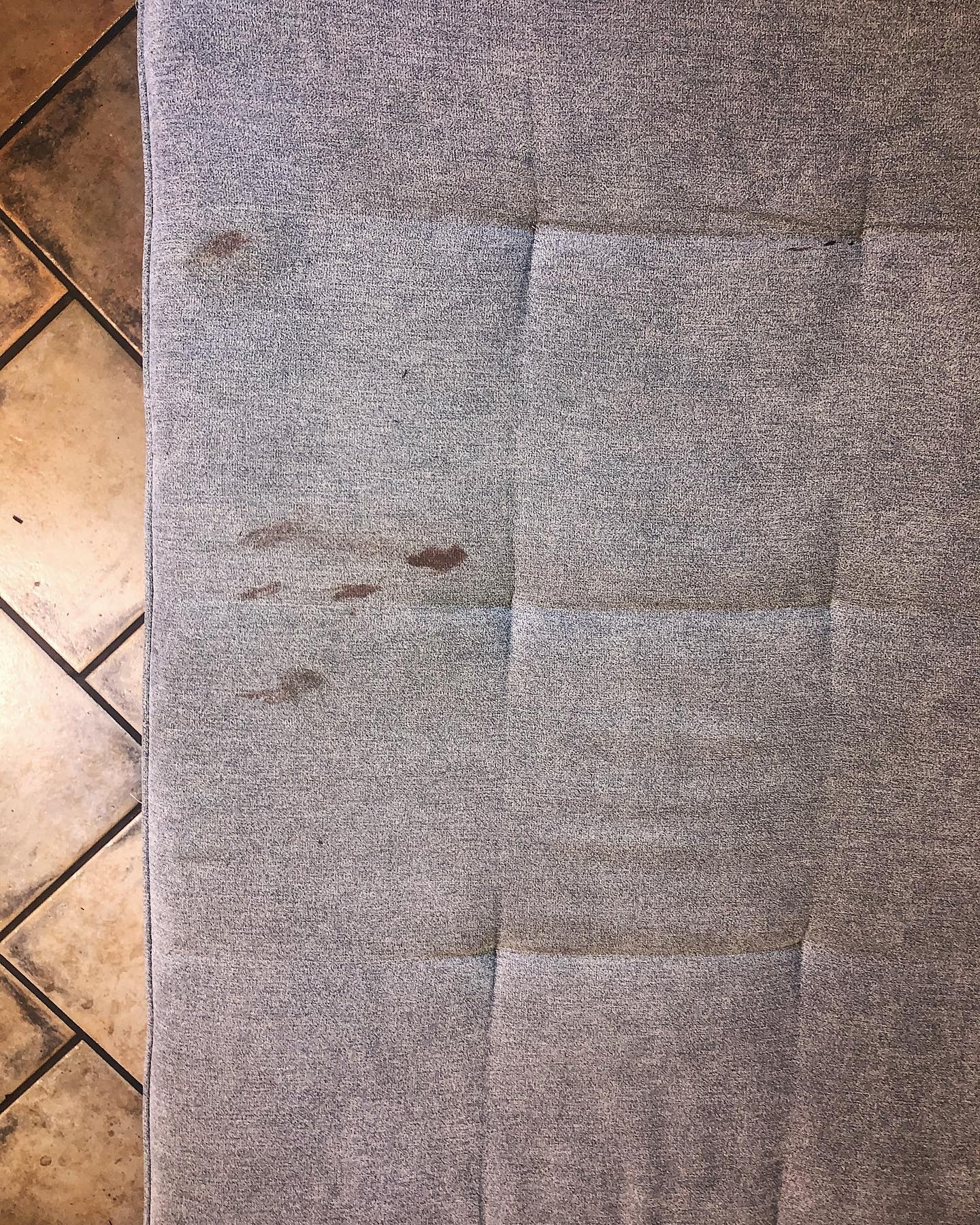 Szara tapicerka mebla z wyraźnymi, ciemnymi plamami po prawej stronie, widoczny fragment podłogi z płytek ceramicznych po lewej stronie.