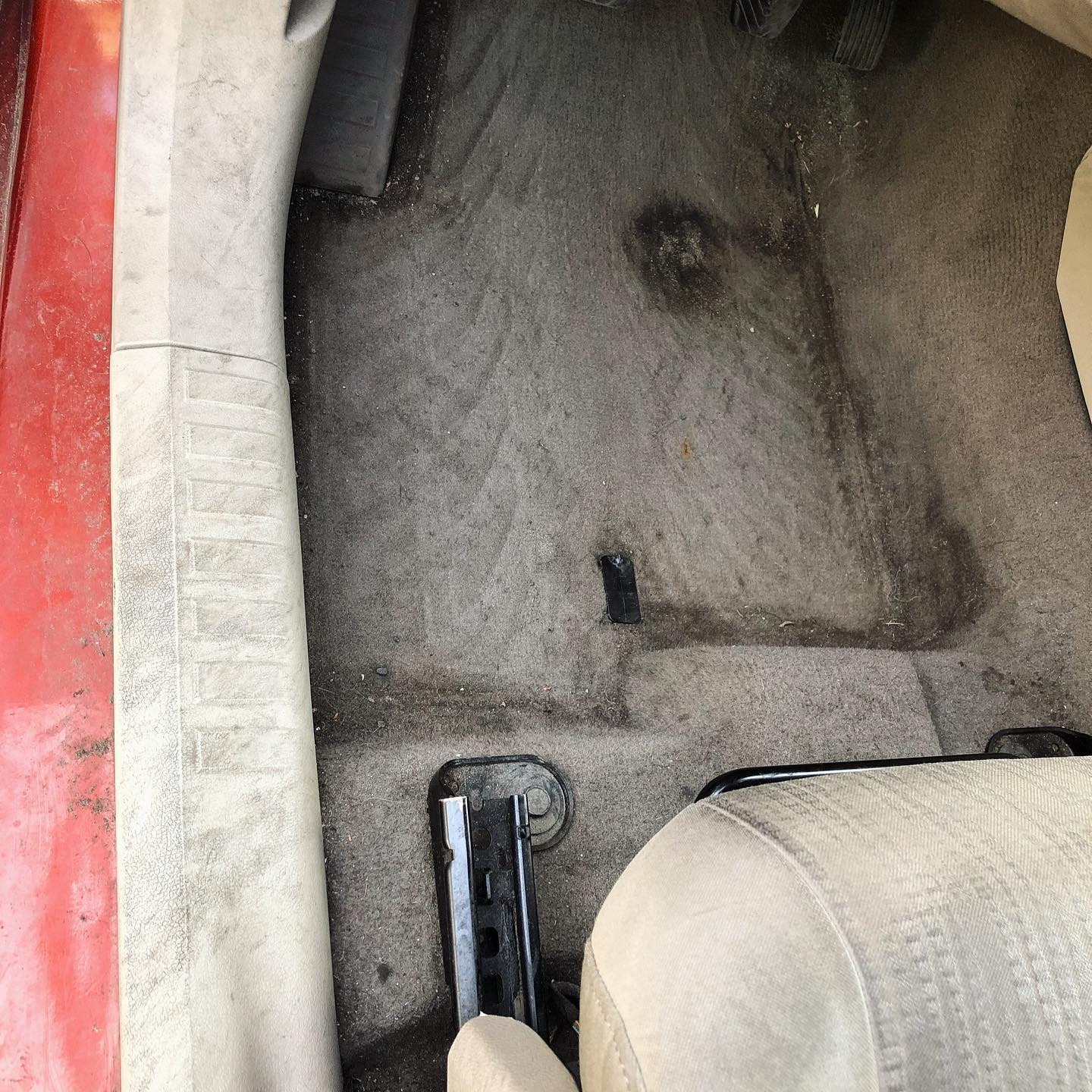 Bardzo zabrudzona, szara wykładzina podłogowa w samochodzie, widoczne plamy i ślady użytkowania, fragment fotela i elementy konstrukcyjne pojazdu.