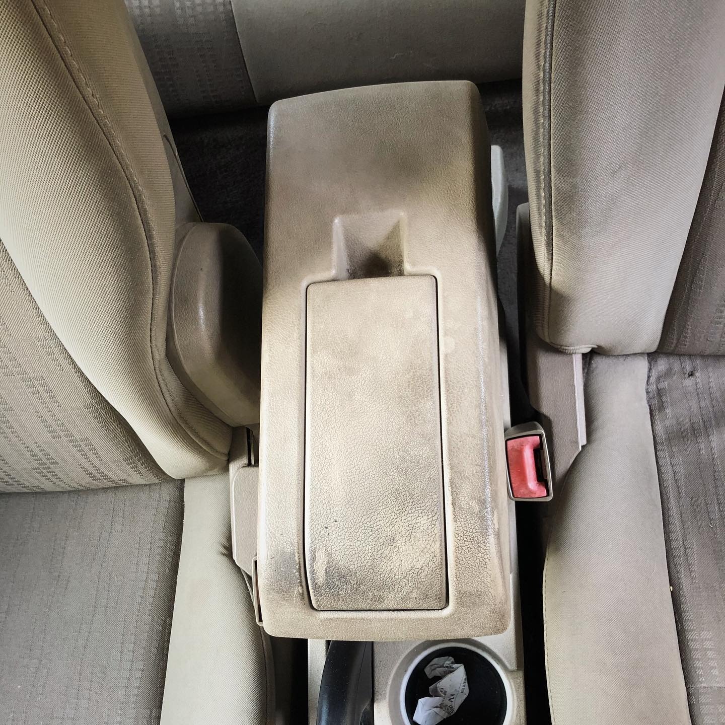 Brudny beżowy podłokietnik samochodowy widziany z góry, z widocznymi plamami i zabrudzeniami, obok fotel pasażera z zapiętym pasem bezpieczeństwa i śmieciami w uchwycie na kubek.