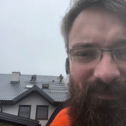 Pruner-Źródło Prądu - Mężczyzna z brodą w pomarańczowej kurtce robi selfie na tle ekipy montującej panele fotowoltaiczne na dachu domu z szarą dachówką.