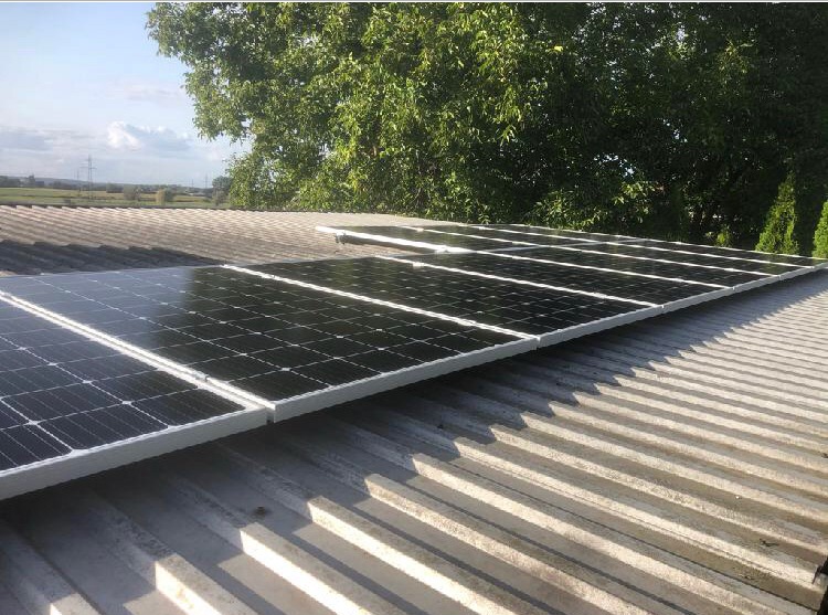 Rząd paneli słonecznych z białymi ramami, zamontowanych na pofałdowanym dachu z blachy, z drzewami i polami w tle.