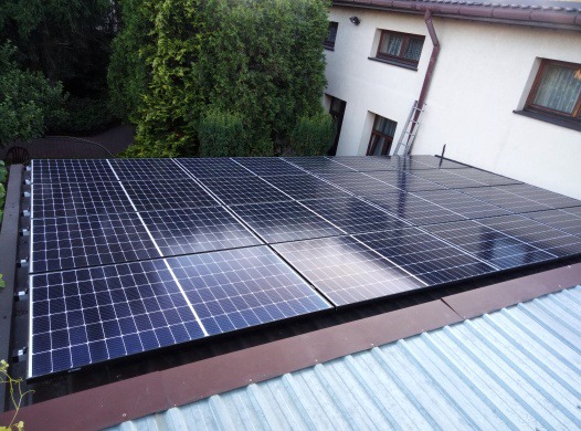 Panele słoneczne zamontowane na metalowym dachu budynku mieszkalnego, widoczne z góry, z drabiną opartą o ścianę w tle.