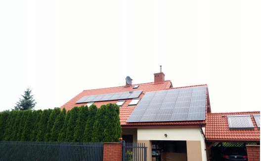 Dom jednorodzinny z czerwoną dachówką, na dachu panele słoneczne i kolektory słoneczne, widok zza żywopłotu.