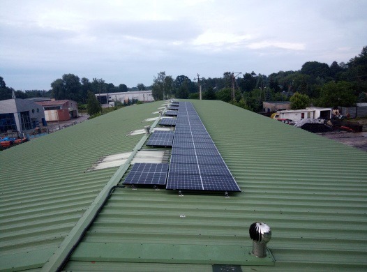 Rząd paneli słonecznych na zielonym, falistym dachu budynku przemysłowego, widok z góry, pochmurne niebo w tle.