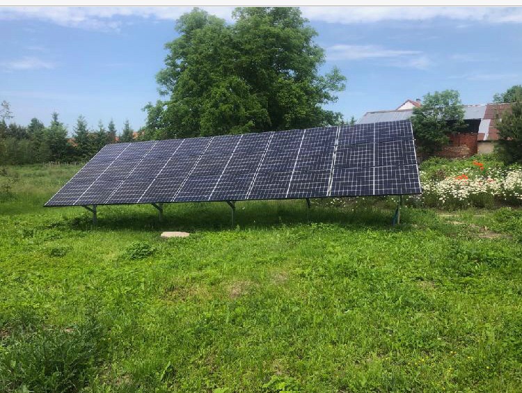 Panel fotowoltaiczny na metalowej konstrukcji, umieszczony na zarośniętej trawie, z widocznymi kwiatami i budynkiem w tle, w słoneczny dzień.