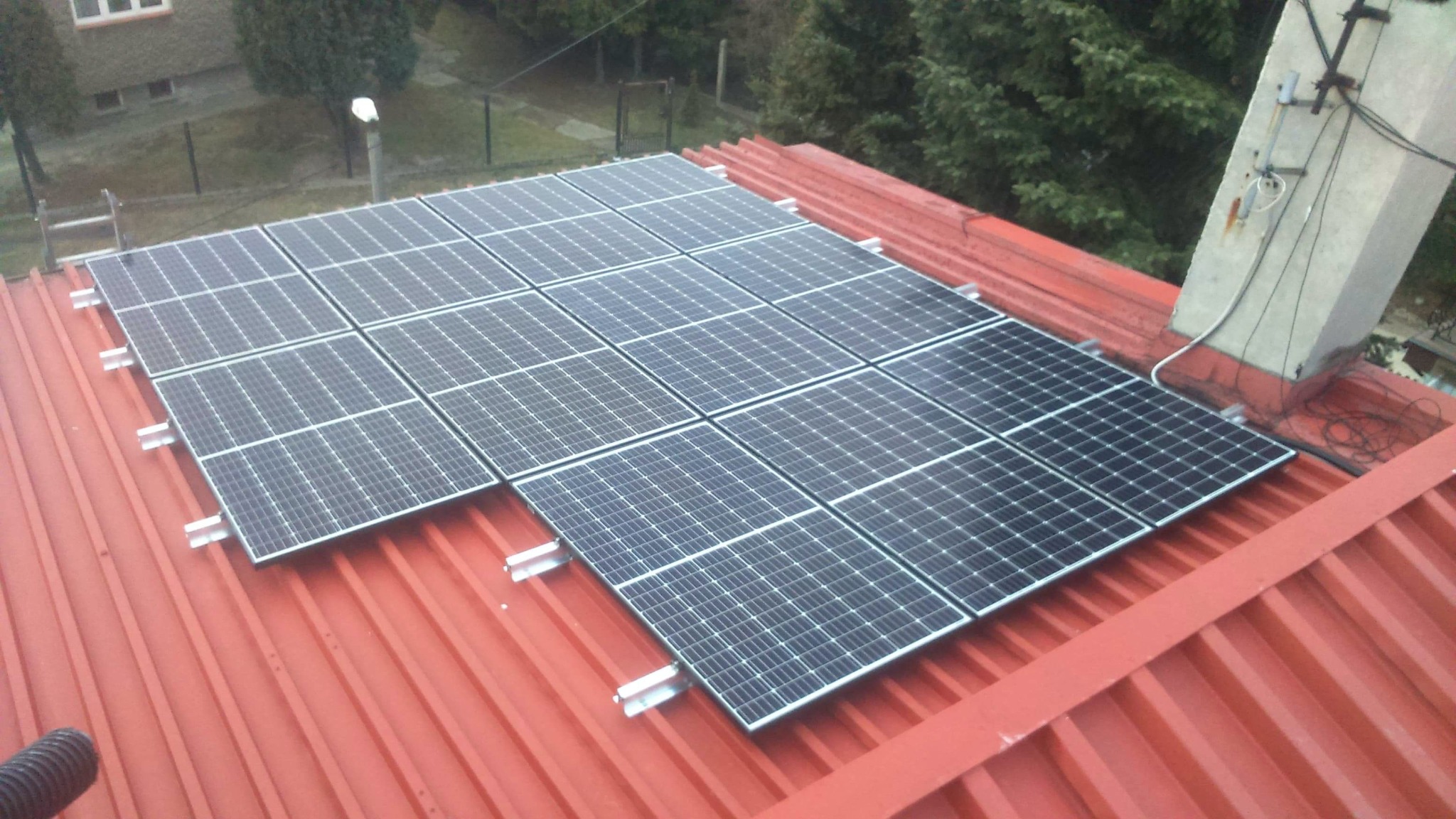 Panele słoneczne zamontowane na czerwonym blaszanym dachu budynku jednorodzinnego, widoczne mocowania i elementy instalacji, w tle drzewa i komin.