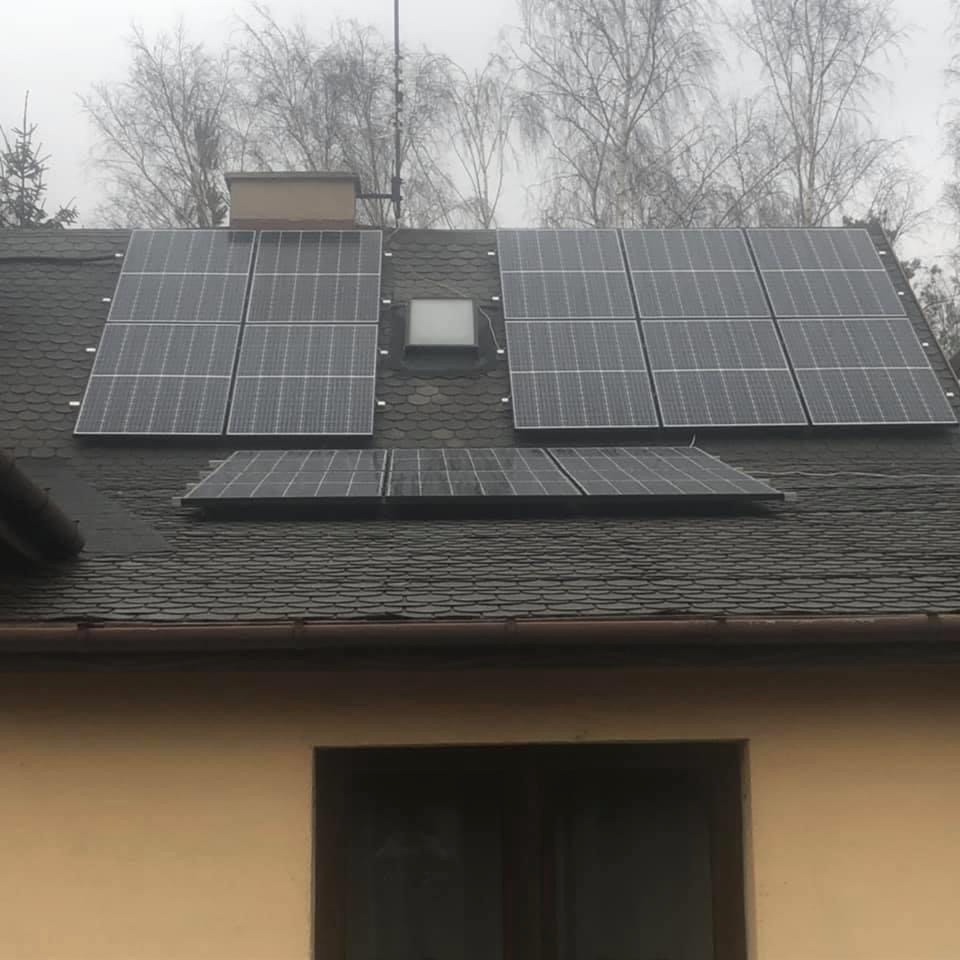 Panele słoneczne zamontowane na dachu pokrytym ciemną dachówką, widoczne okno dachowe i fragment elewacji budynku w pochmurny dzień.