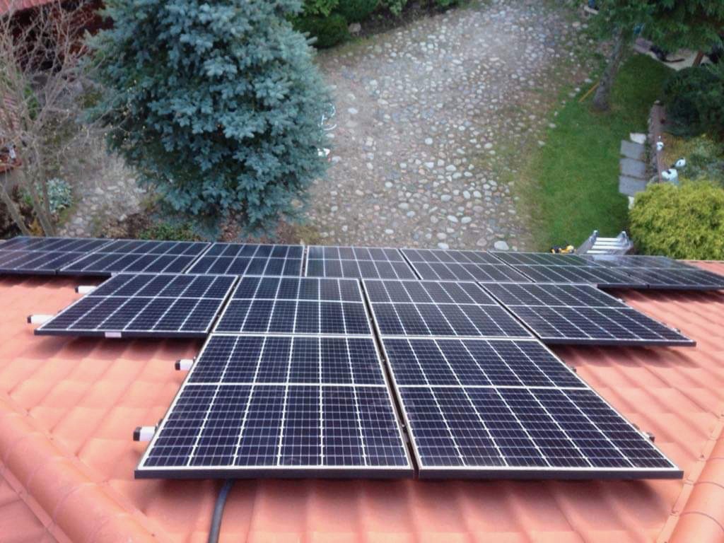 Instalacja paneli słonecznych na pomarańczowym dachu z widocznymi elementami montażowymi i otoczeniem domu: drzewa iglaste, ścieżka z kamienia, trawnik.