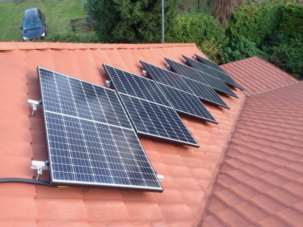 Rząd paneli słonecznych zamontowanych na czerwonym, ceramicznym dachu domu jednorodzinnego, widok z góry, w tle zaparkowany samochód.