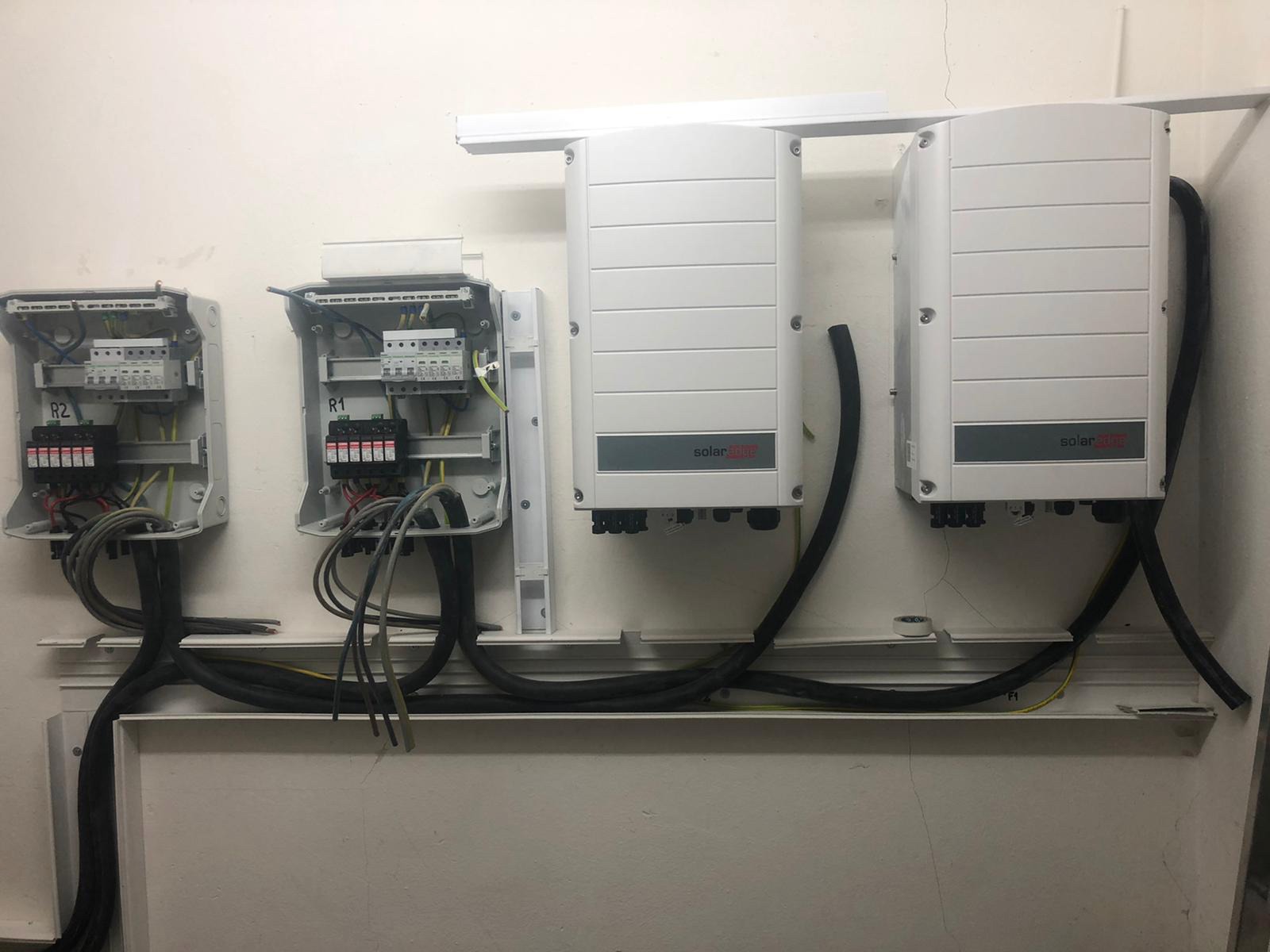 Dwa białe inwertery SolarEdge zamontowane na ścianie obok dwóch otwartych skrzynek elektrycznych z okablowaniem i bezpiecznikami, czarne kable prowadzone wzdłuż ściany.