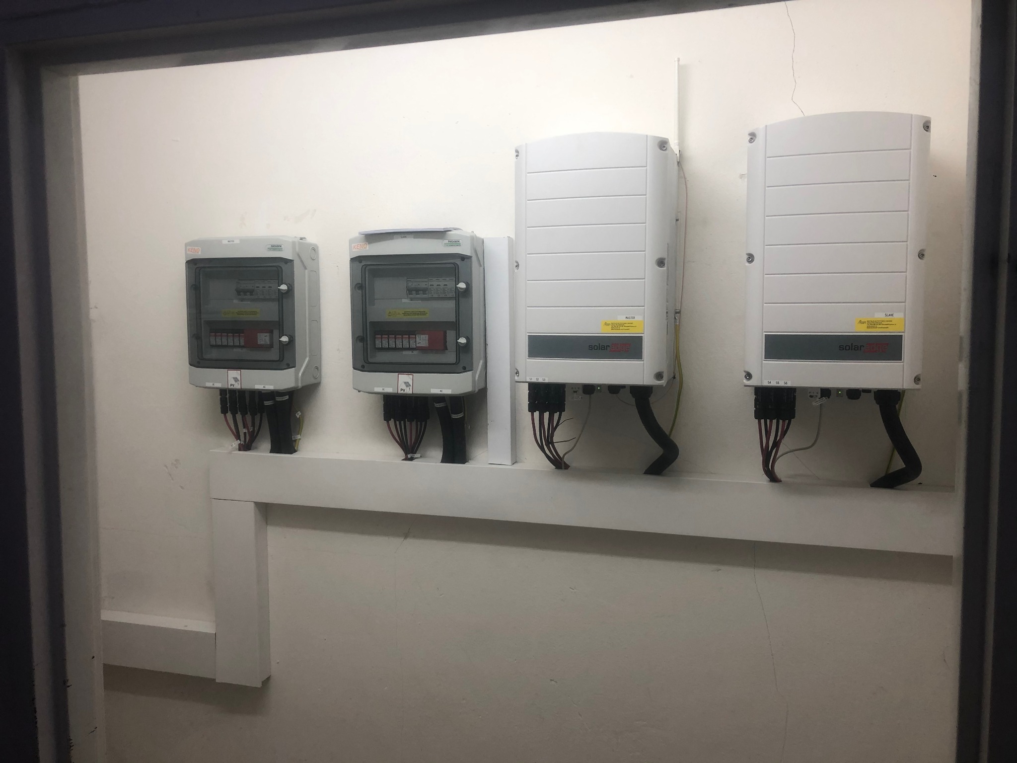 Dwa falowniki SolarEdge i dwie skrzynki zabezpieczeń fotowoltaicznych zamontowane na białej ścianie i wsporniku, widoczne kable połączeniowe.