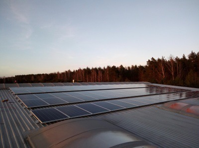 Panele słoneczne zamontowane na falistym dachu przemysłowym z widokiem na las w oddali, oświetlone ciepłym światłem zachodzącego słońca.