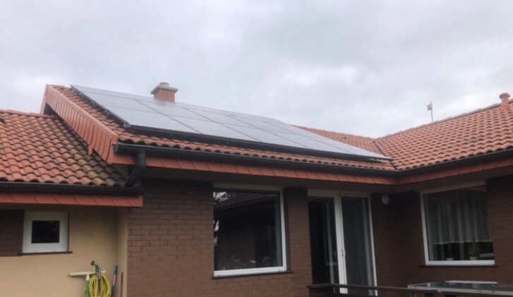 Panele słoneczne zamontowane na dachu domu z czerwoną dachówką, widoczne okna i ceglana ściana domu, pochmurne niebo.