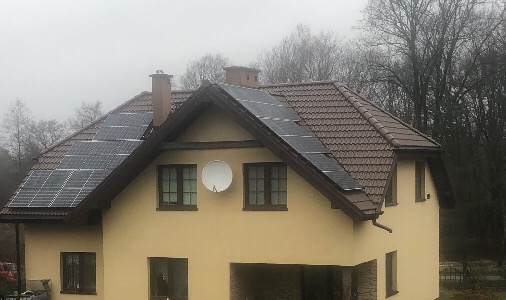 Instalacja fotowoltaiczna na brązowym dachu dwuspadowym domu jednorodzinnego z kremową elewacją i anteną satelitarną, widok z poziomu gruntu w pochmurny dzień.